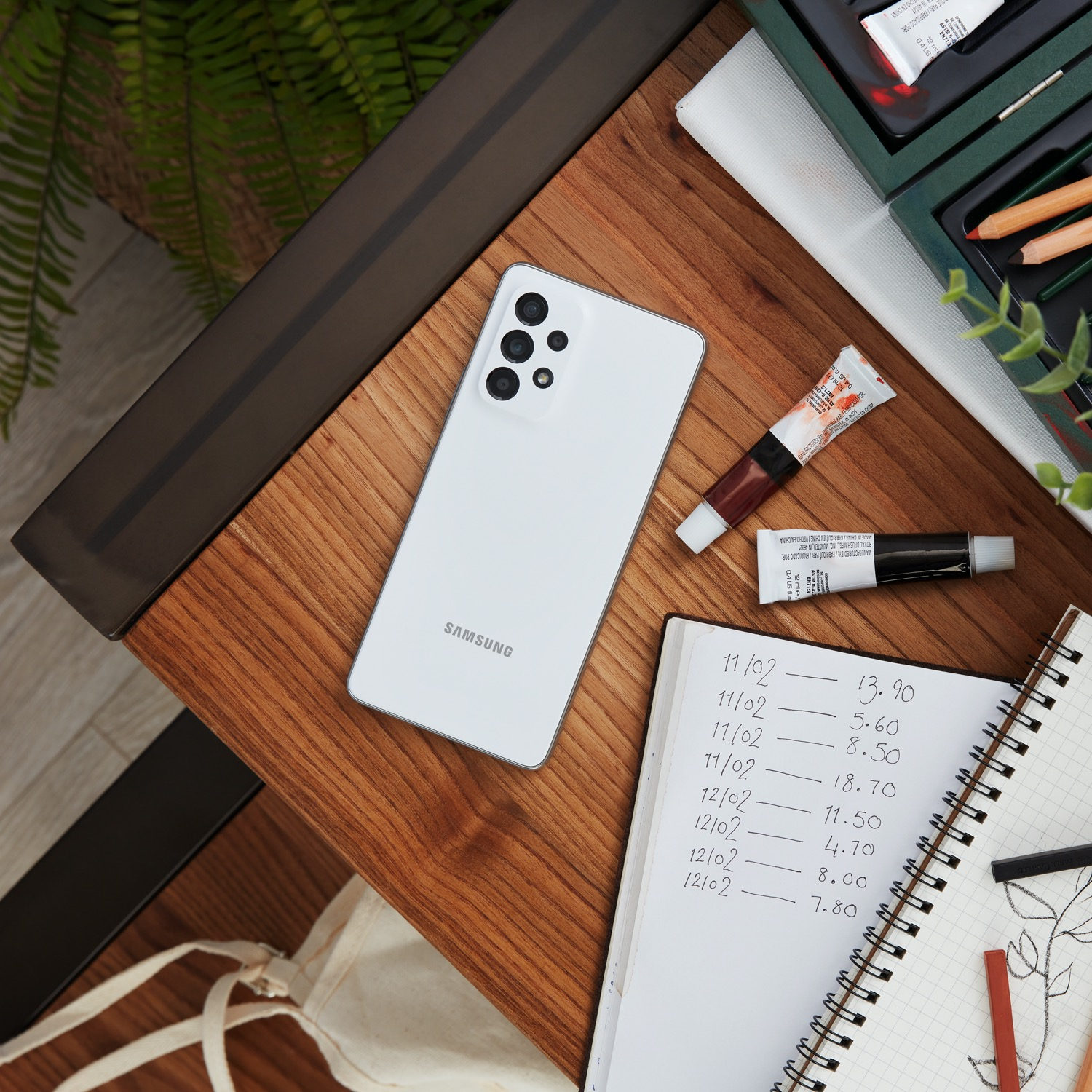 Teléfono Samsung blanco, cuaderno abierto con notas, tubos de pintura y útiles de arte sobre mesa madera.