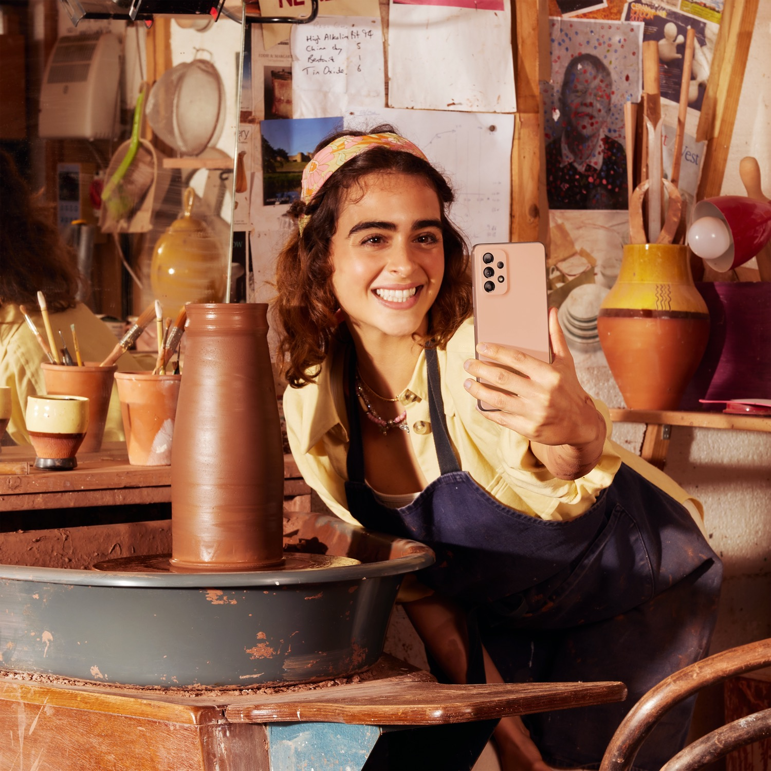 Mujer sonriendo, sosteniendo un teléfono rosa, en un estudio de cerámica. Fondo con cerámica y herramientas.