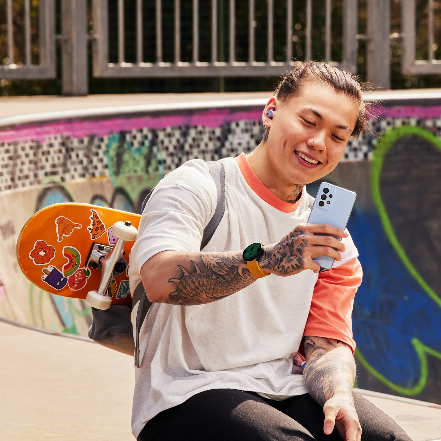 Joven con tatuajes sonríe, sosteniendo un teléfono azul y usando auriculares, patinando en el skatepark.