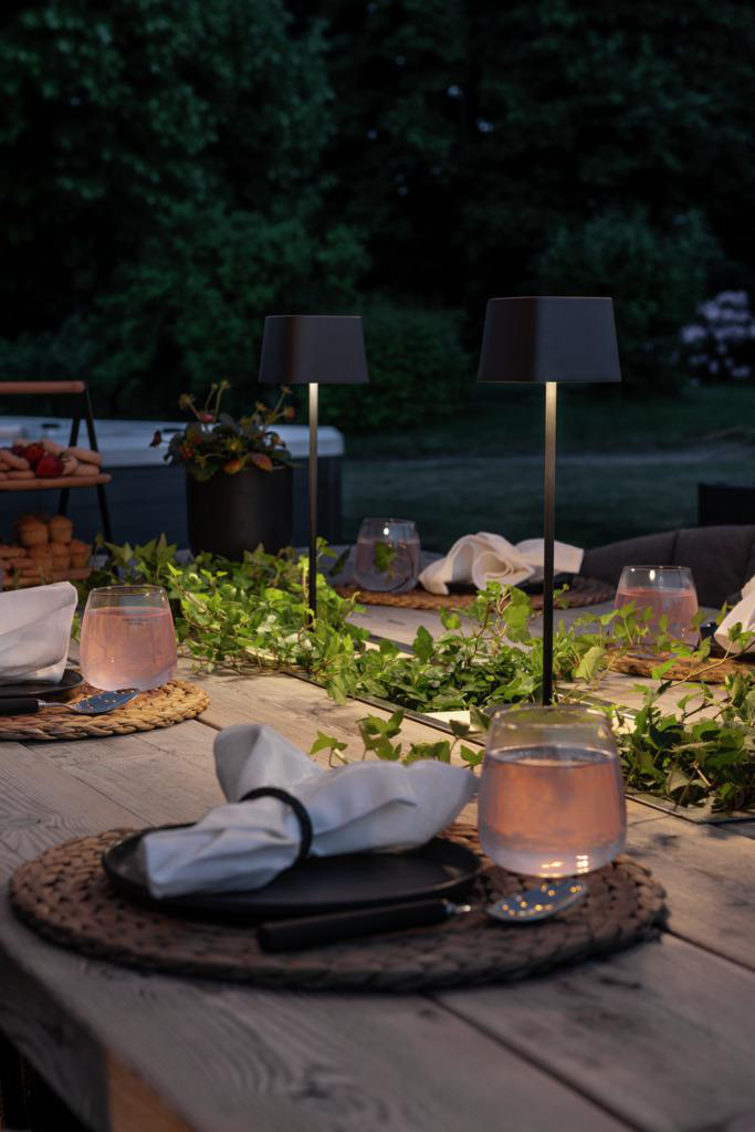 Draußen gedeckter Tisch bei Dämmerung mit Lampen, Gläsern und Grün.