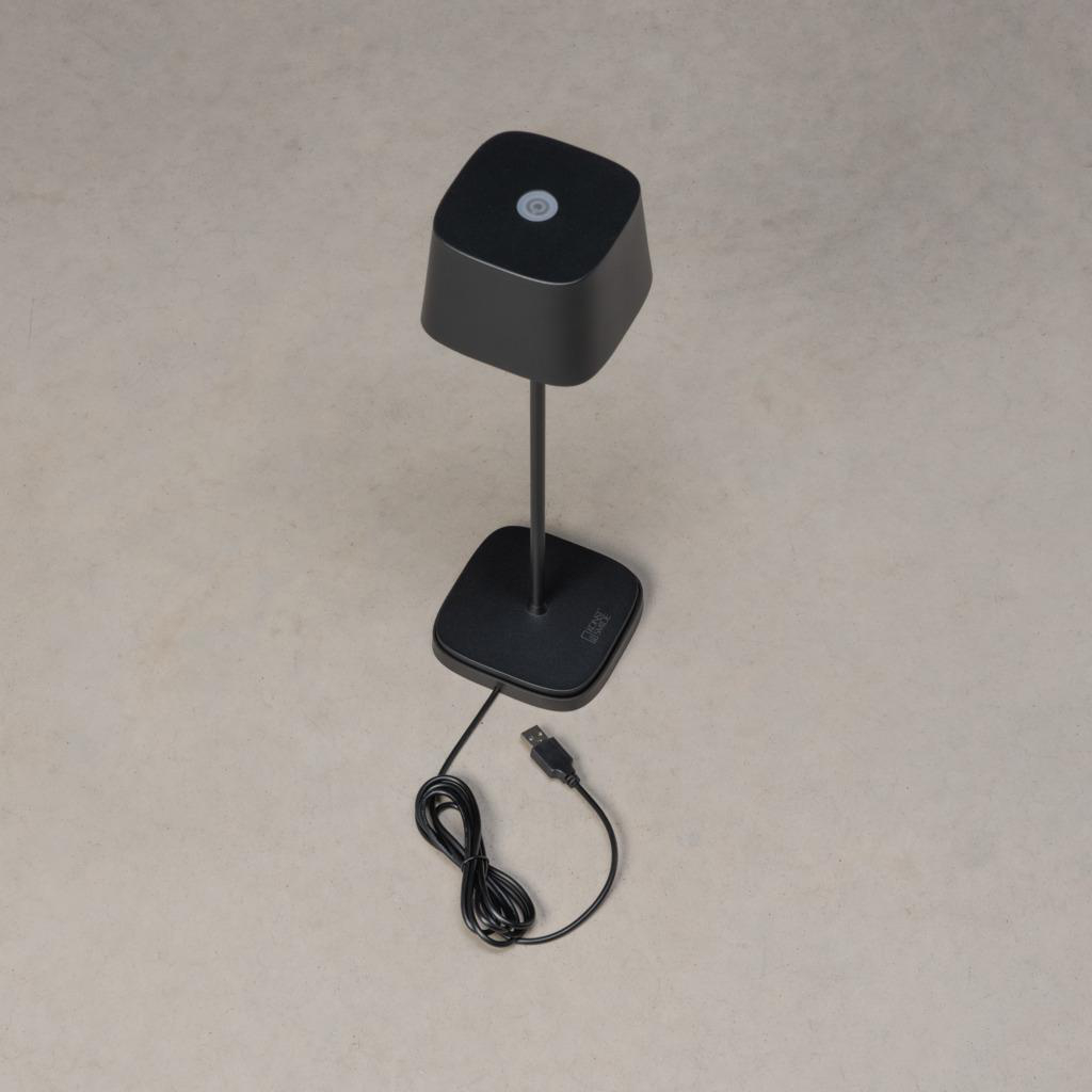 Schwarze würfelförmige Lampe auf einem schwarzen Ständer. Ein Kabel und USB-Stecker sind befestigt. Hellbeiger Hintergrund.