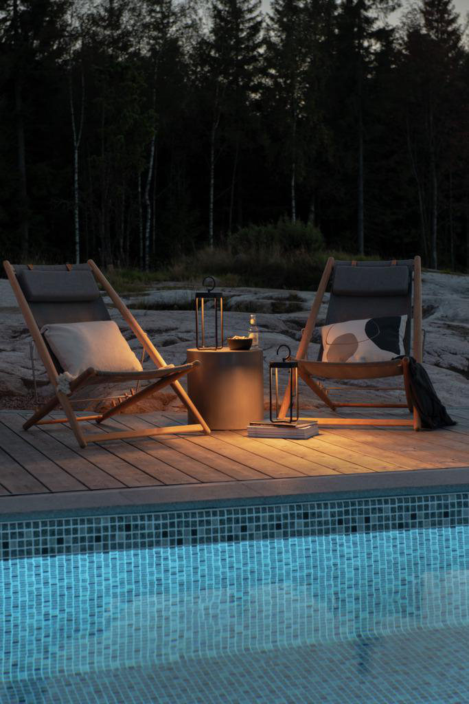 Zwei hölzerne Liegestühle und ein kleiner Tisch an einem Pool in der Dämmerung. Laternen spenden Licht.