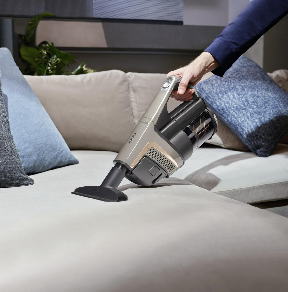 Hand, die einen Staubsauger über einem hellfarbigen Sofa hält. Blaue Kissen im Hintergrund.