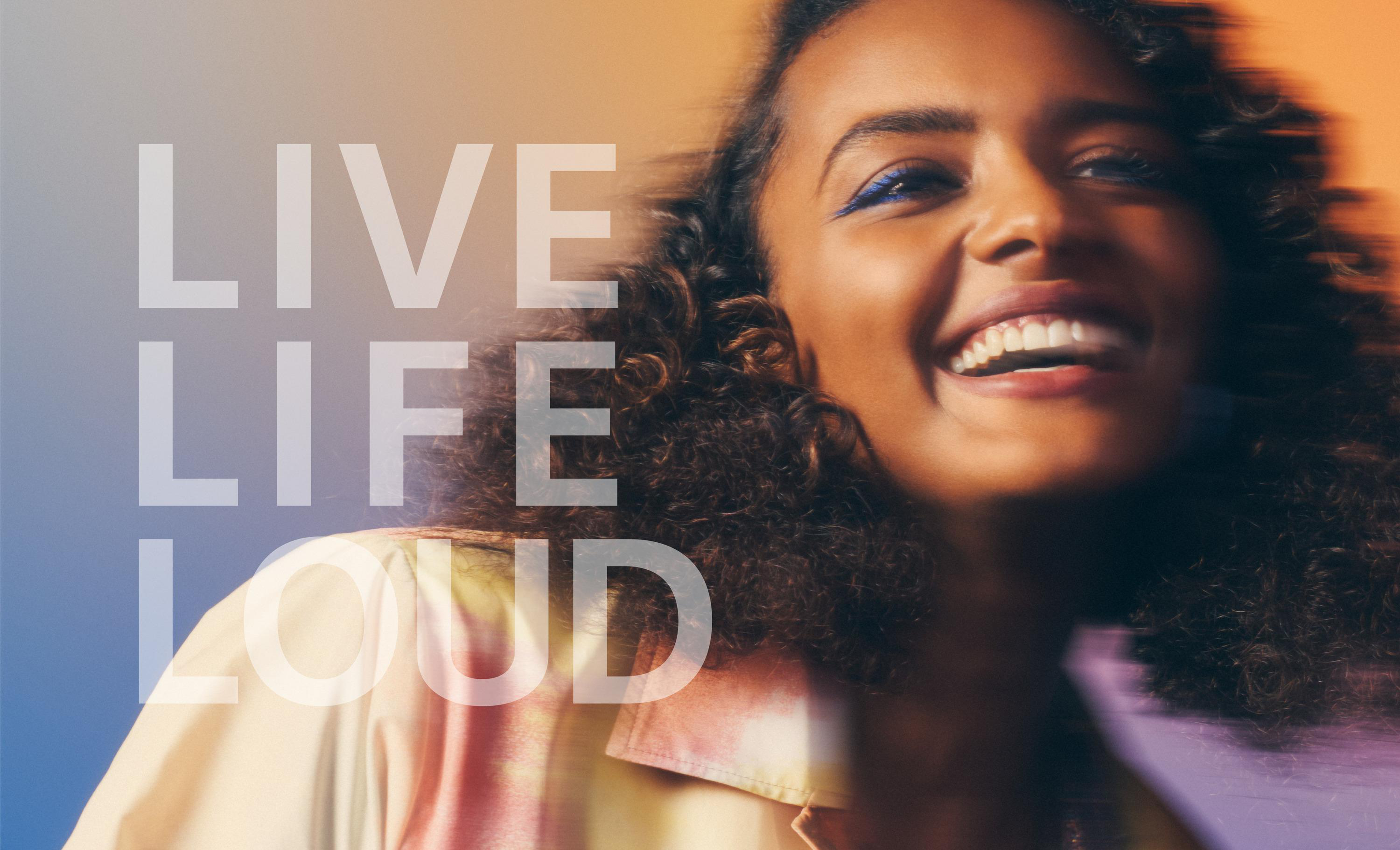 Frau lächelt, trägt farbenfrohes Hemd. Text „Live Life Loud“. Verschwommener Hintergrund.