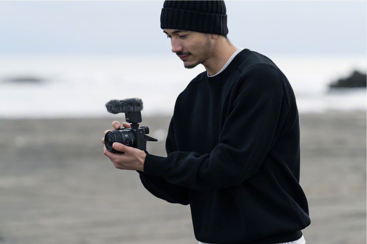 Mann in schwarzem Pullover und Mütze hält Kamera mit Mikrofon, Strand im Hintergrund.