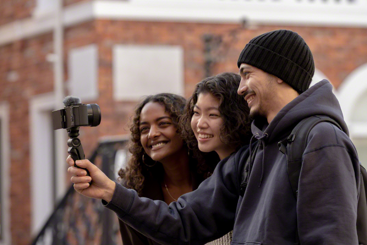 Drei lächelnde Personen machen ein Selfie mit einer Kamera, mit einem Backsteingebäude im Hintergrund.