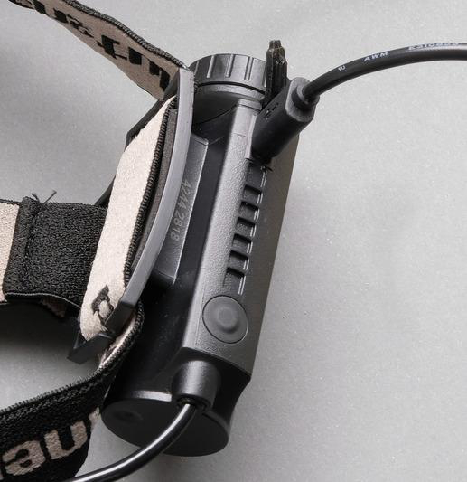 Schwarze Stirnlampe mit Gurt und Ladeanschluss. Die Stirnlampe ist abgewinkelt.
