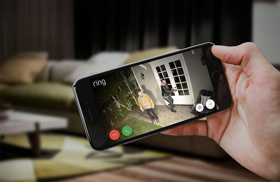 A person holding a phone displaying a Ring app view of a front door with people and a yard.