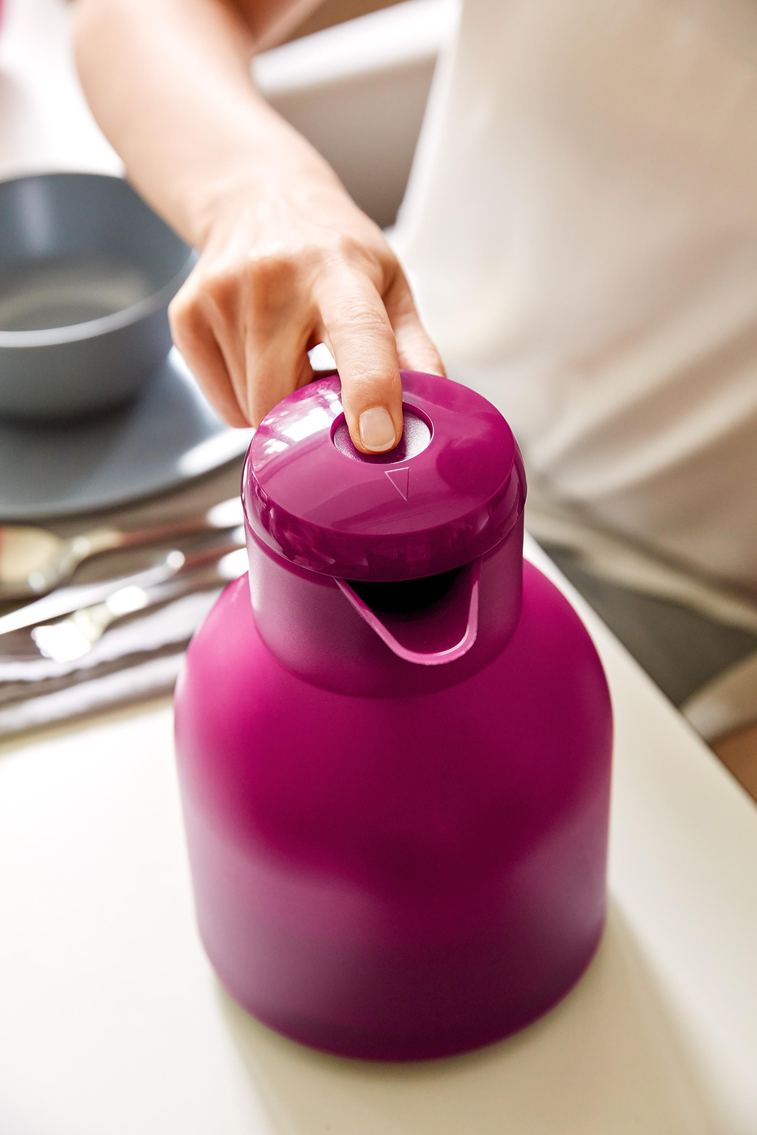 Hand drückt den magenta Deckel einer Thermoskanne. Eine Schüssel und Besteck sind im unscharfen Hintergrund.