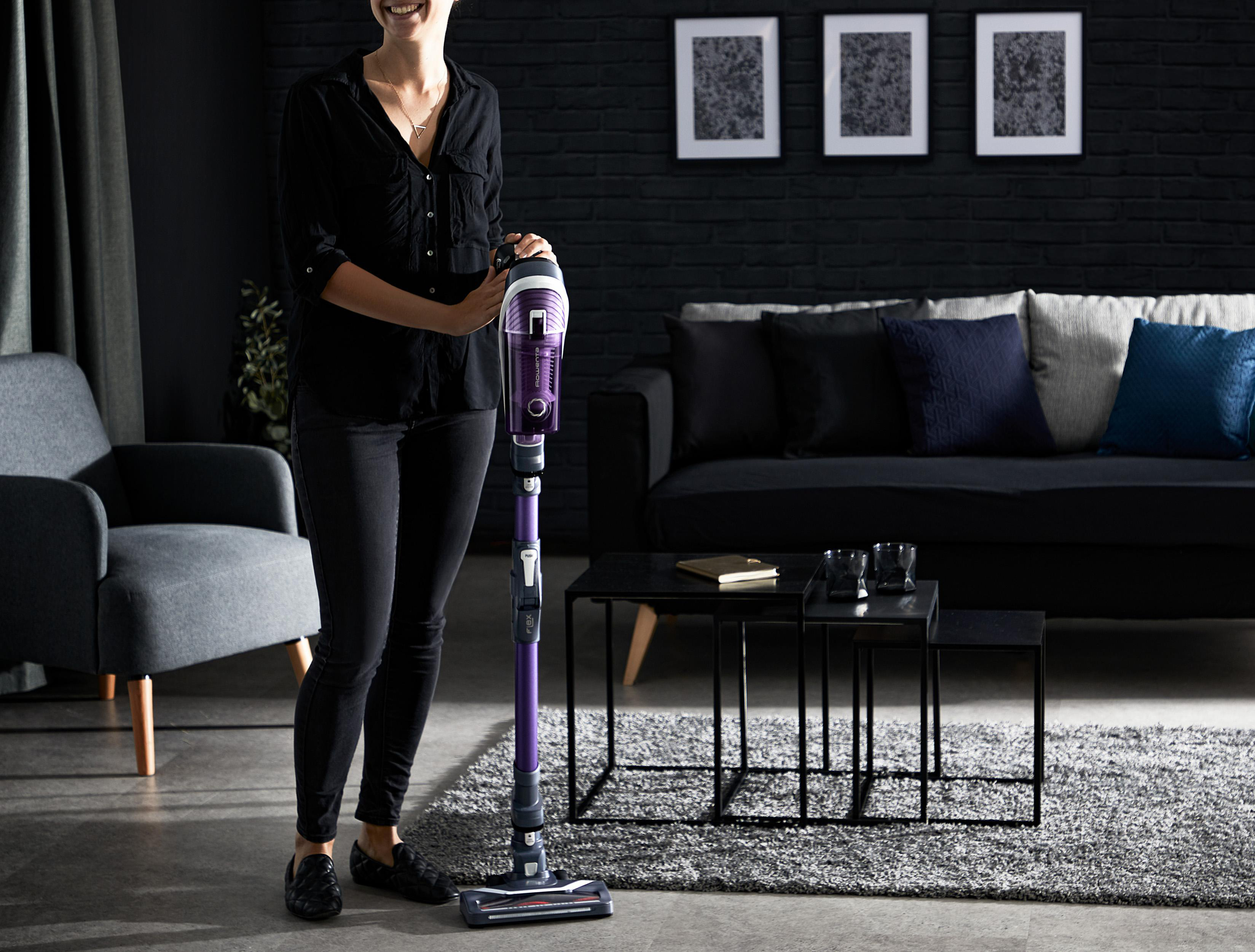 Lächelnde Frau, die einen lila Staubsauger in einem modernen Wohnzimmer hält.
