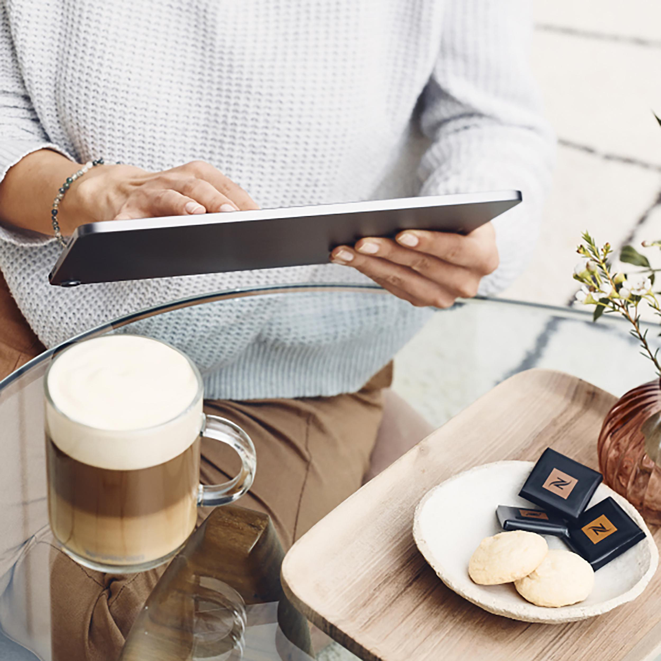 Eine Person hält ein Tablet, eine Kaffeetasse und einen Teller mit Keksen auf einem Tisch.