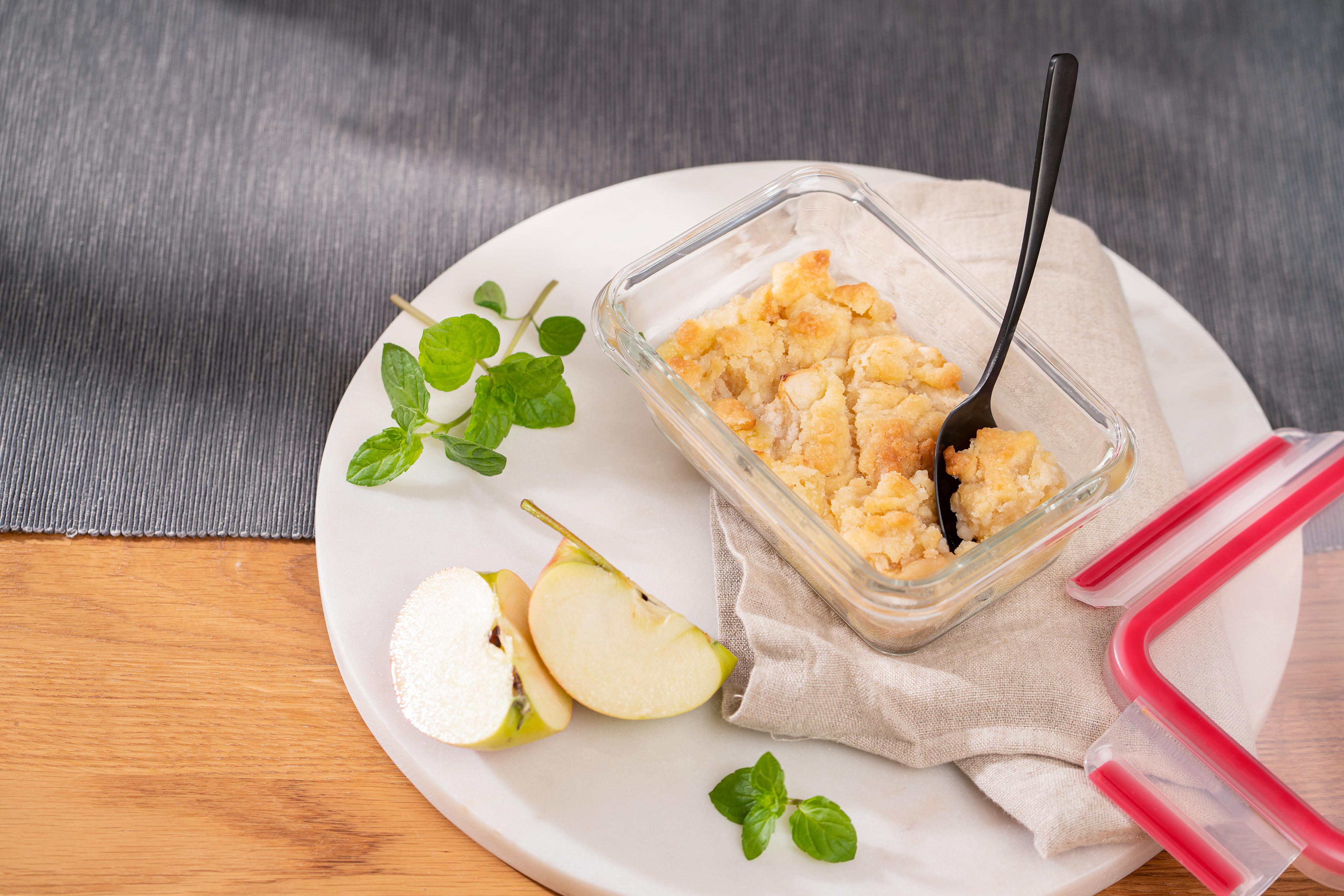 Apfel-Crumble-Dessert in einer Glasschale auf einem weißen Teller, mit Apfelscheiben und Minze.