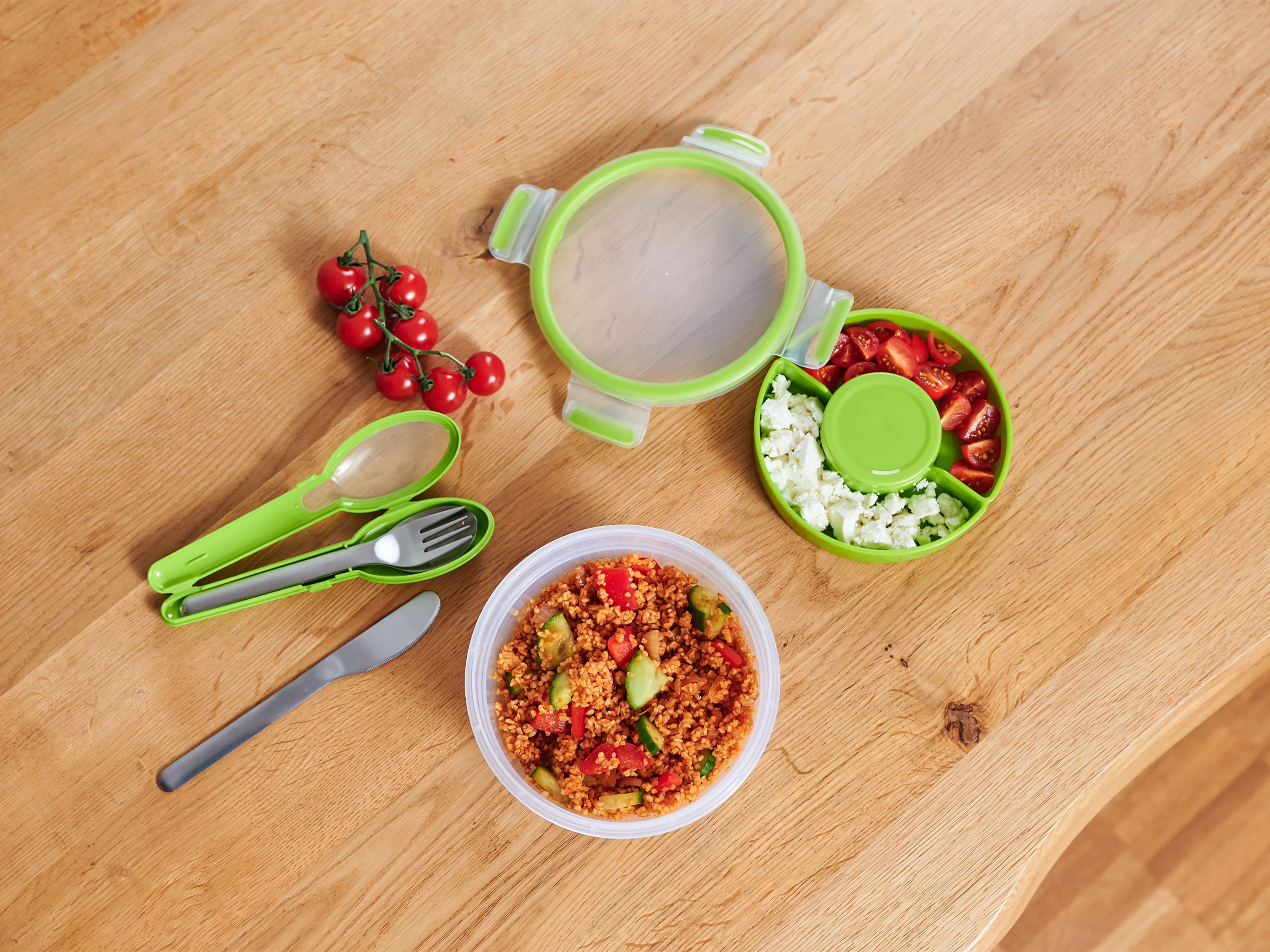 Lunchbox mit Fächern mit Salat, Couscous und Tomaten auf einem Holztisch.