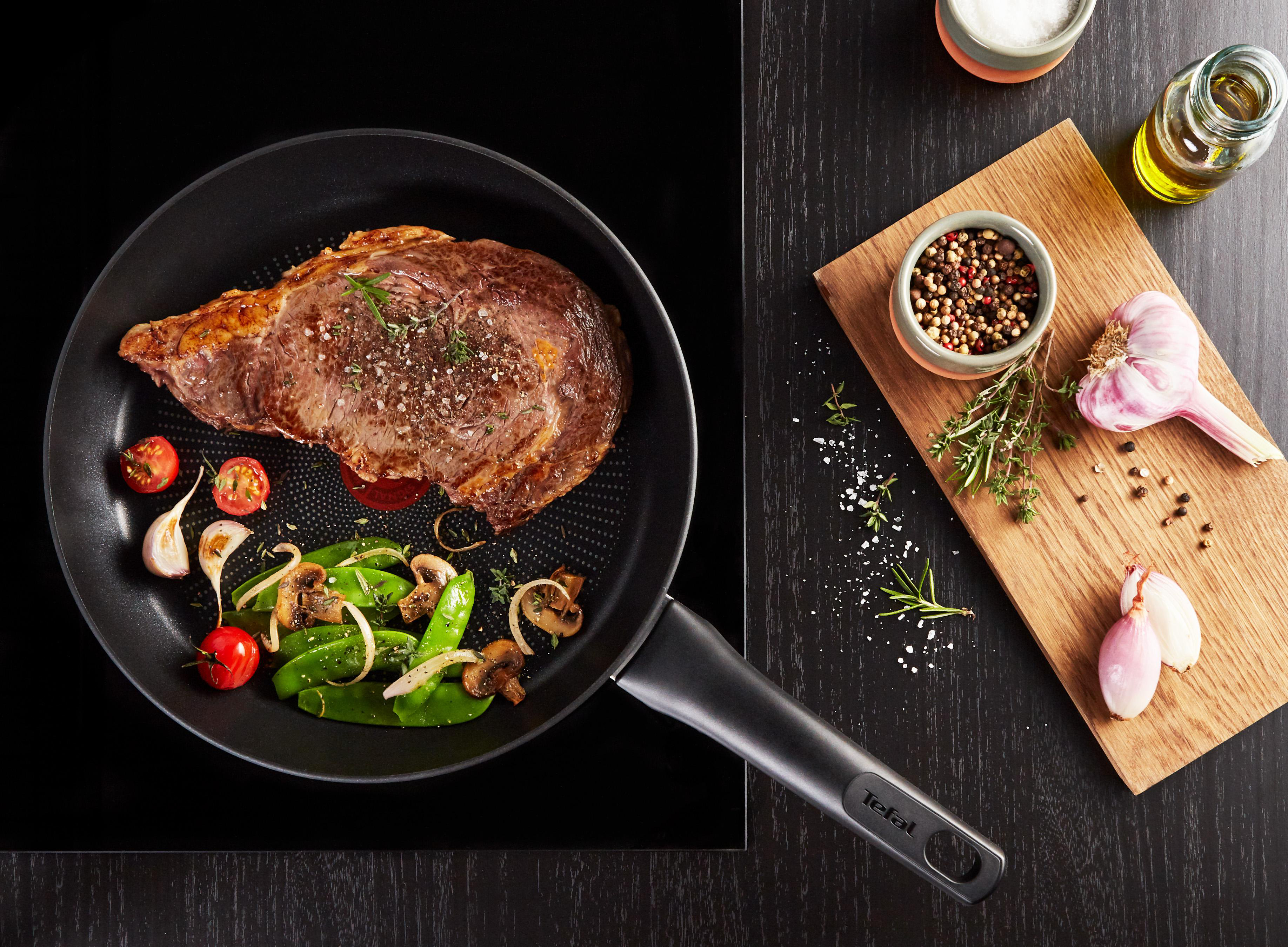 Steak in der Pfanne mit Gemüse und Knoblauch auf Holzbrett, auf schwarzer Oberfläche.