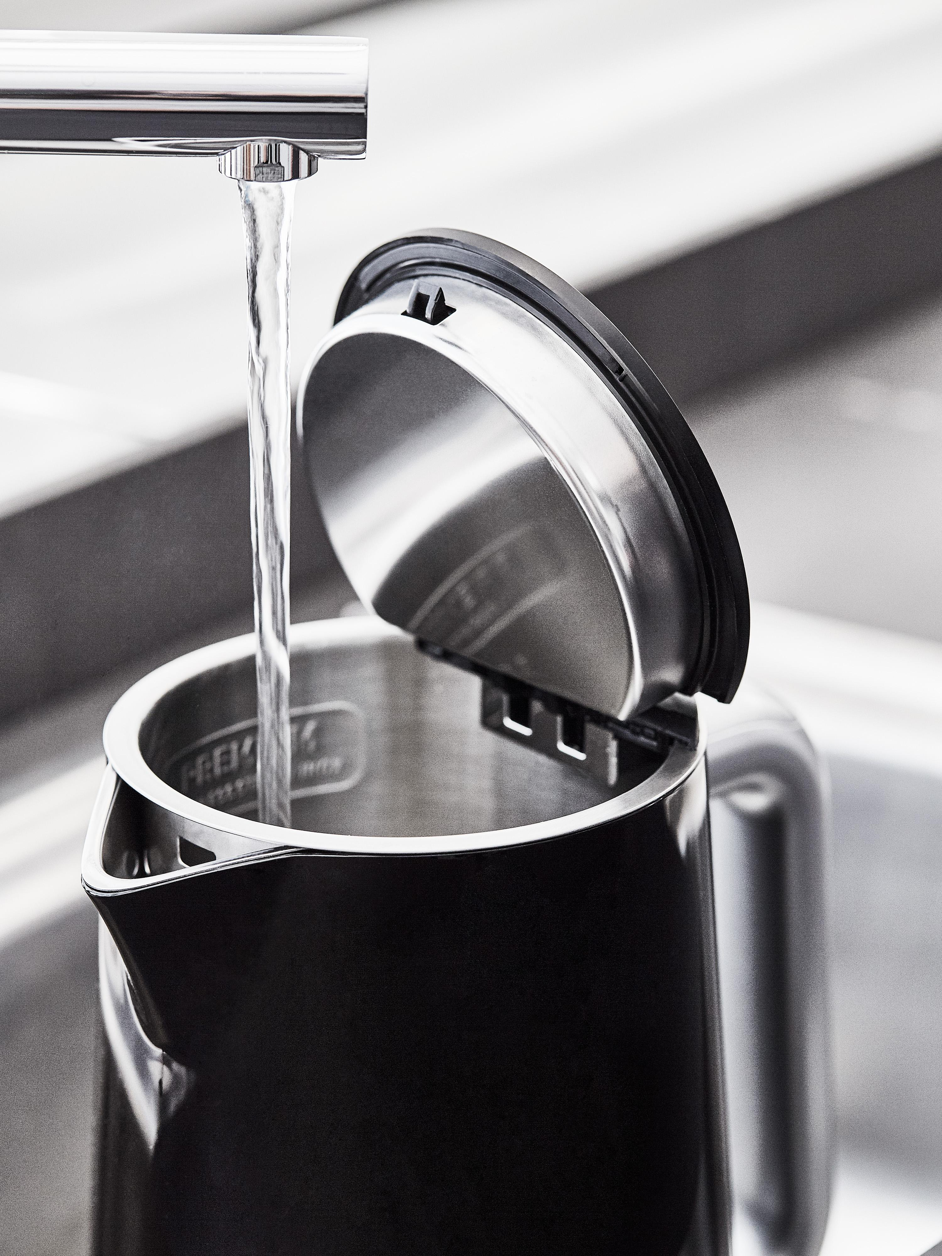 Wasser gießt in einen schwarzen Wasserkocher mit offenem Deckel, Nahaufnahme.