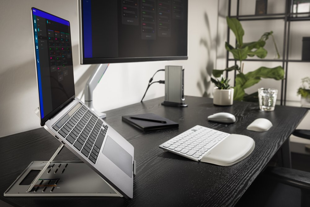 Laptop állványon, monitor, billentyűzet, egér és íróasztali elemek sötét asztalon.