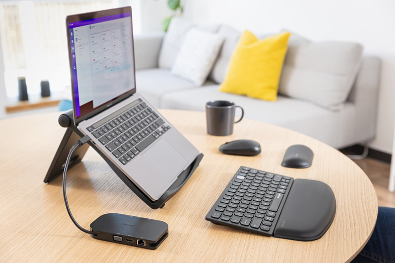 Laptop állványon, billentyűzet, egér, bögre kerek asztalon. Kanapé a háttérben.