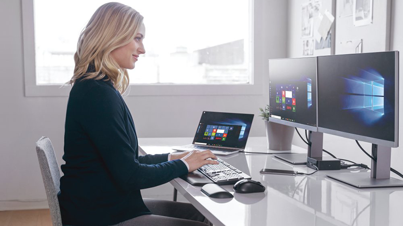 Nő dolgozik egy asztalnál, laptop, billentyűzet, két monitor és egér.