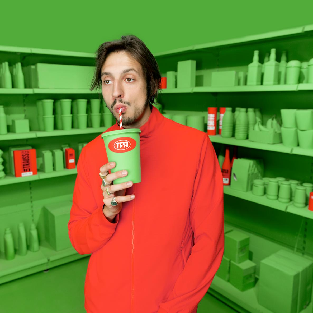 Homme buvant dans un gobelet vert, veste rouge, dans magasin vert.