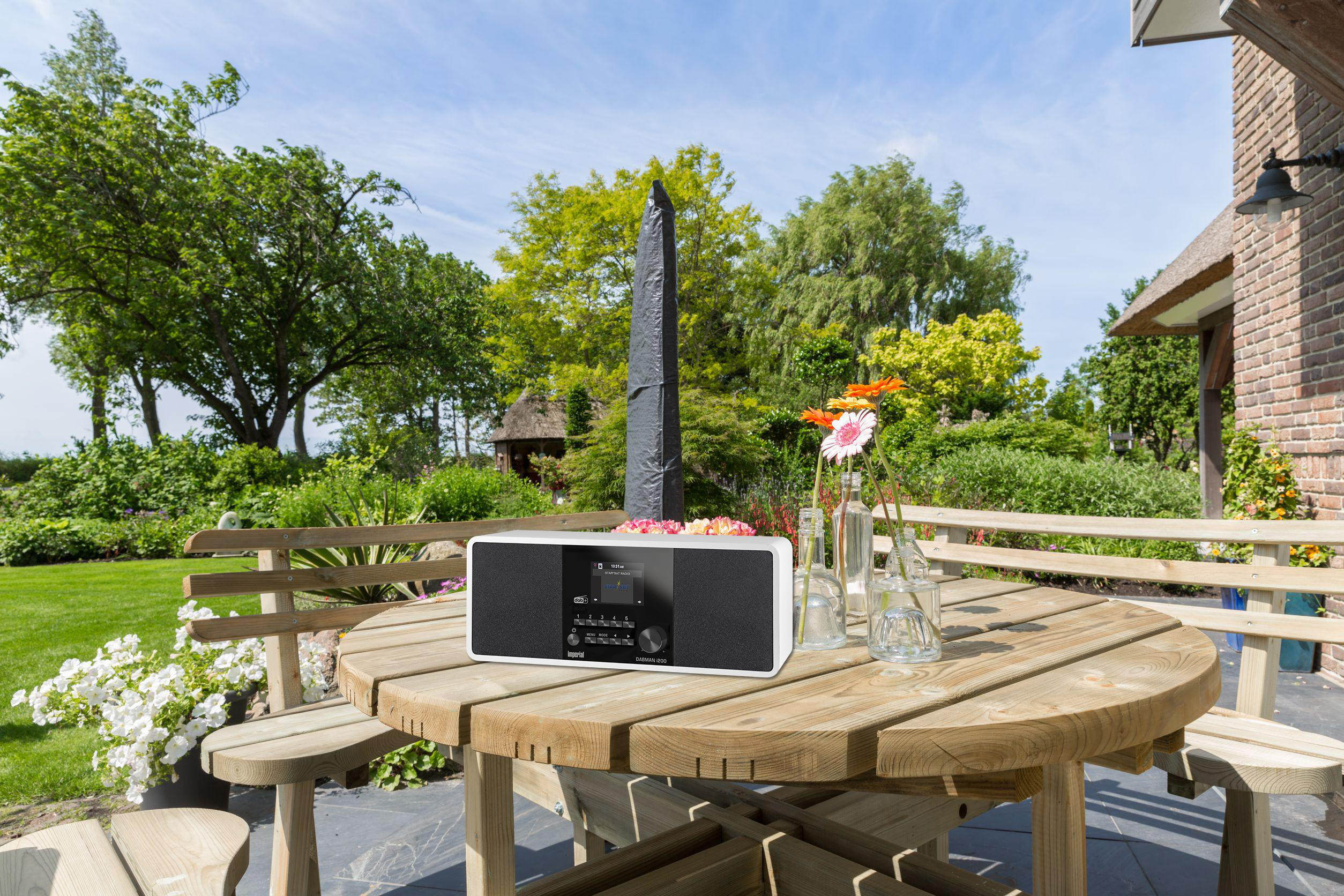 Ein Radio steht draußen auf einem Holztisch mit Blumen und Garten im Hintergrund.