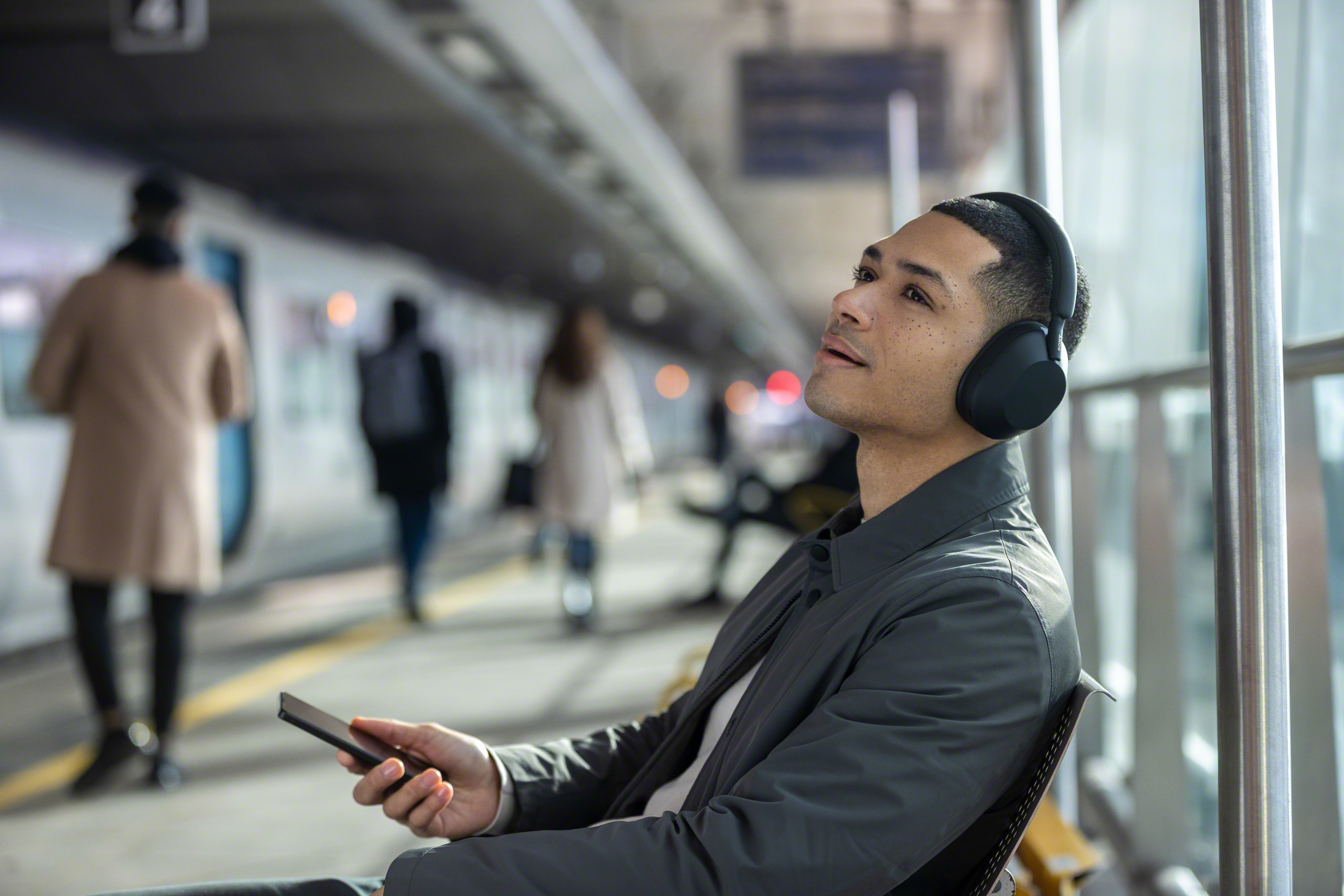 Siyah kulaklık takan bir adam, bir tren istasyonunda yukarı bakıyor, telefon tutuyor. Arka planda insanlar ve bir tren var.