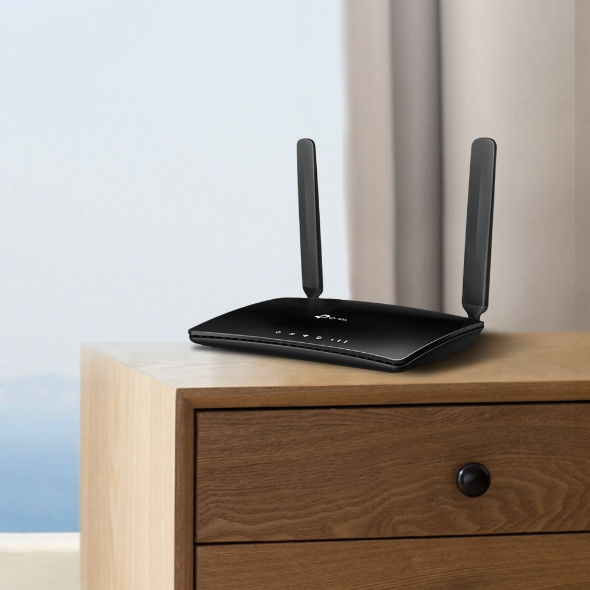 Black router on a wooden surface, with two antennas pointing upwards. Background: window and curtains.