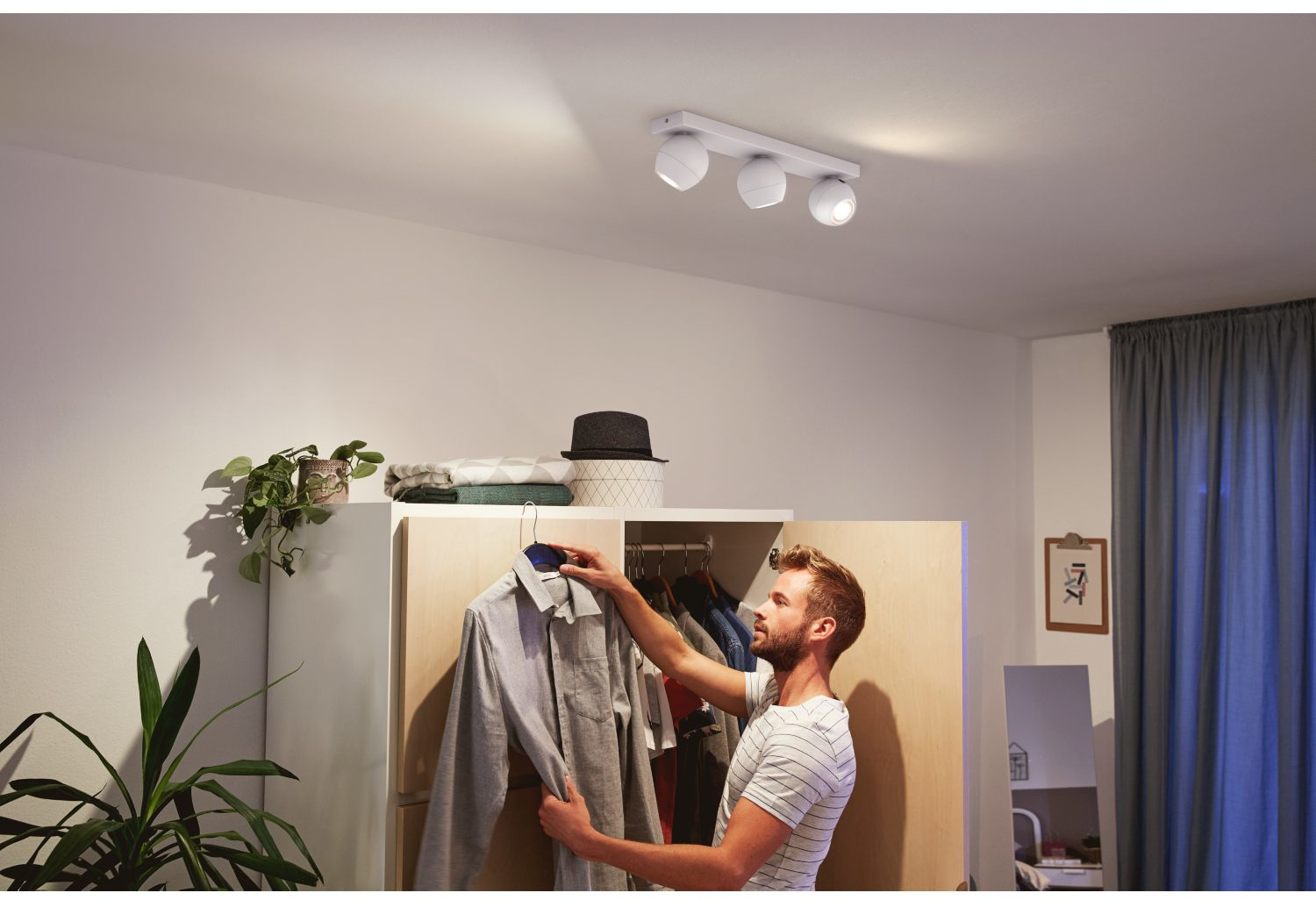 Een man die naar een overhemd reikt in een open kast, onder drie witte spots aan het plafond.
