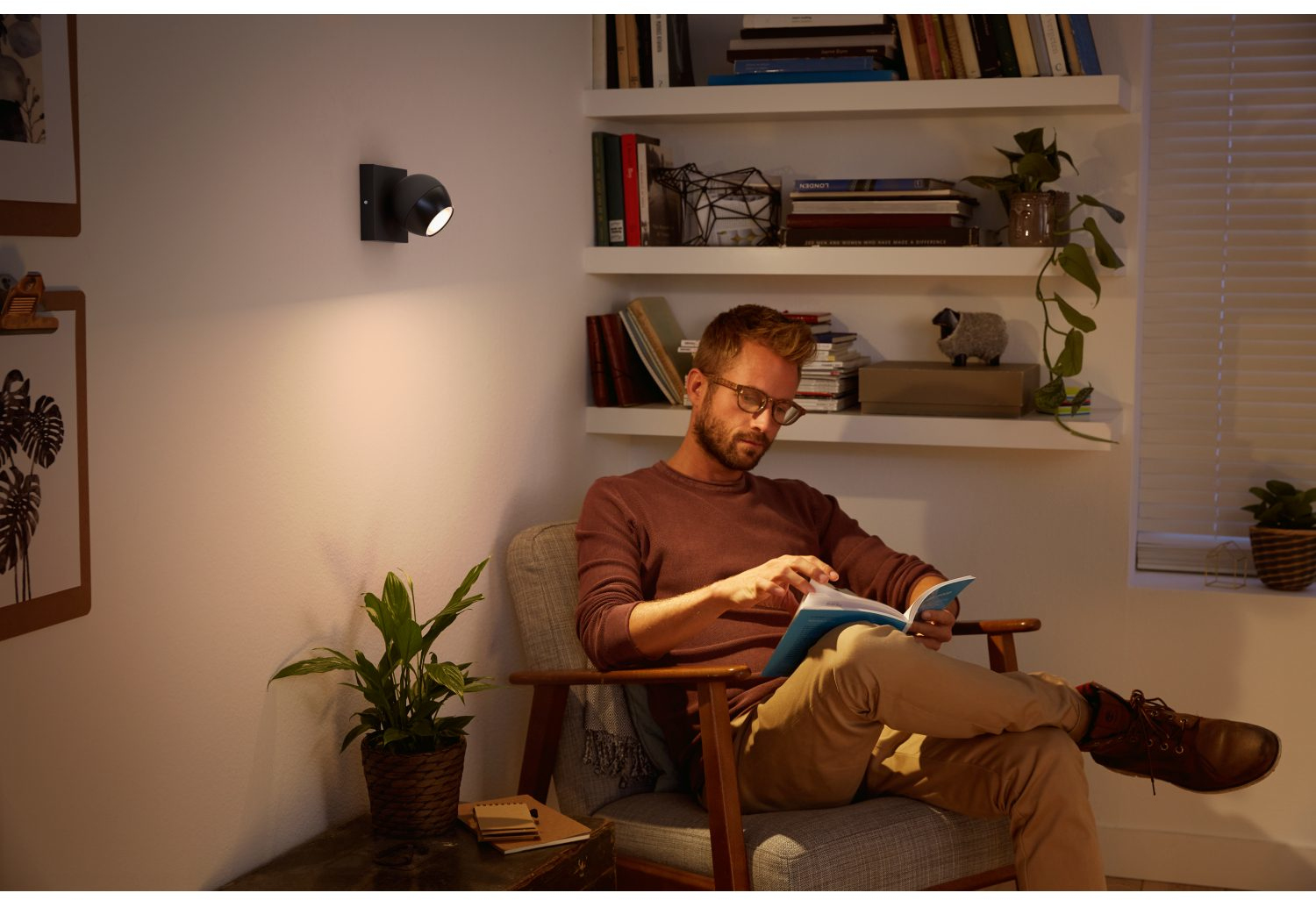 Een man met een bril zit in een stoel en leest een boek met een wandlamp.
