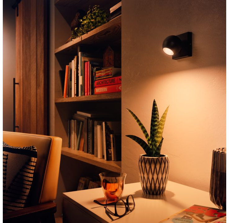 Woonkamer met boekenplank, tafel met plant en bril, en wandlamp.