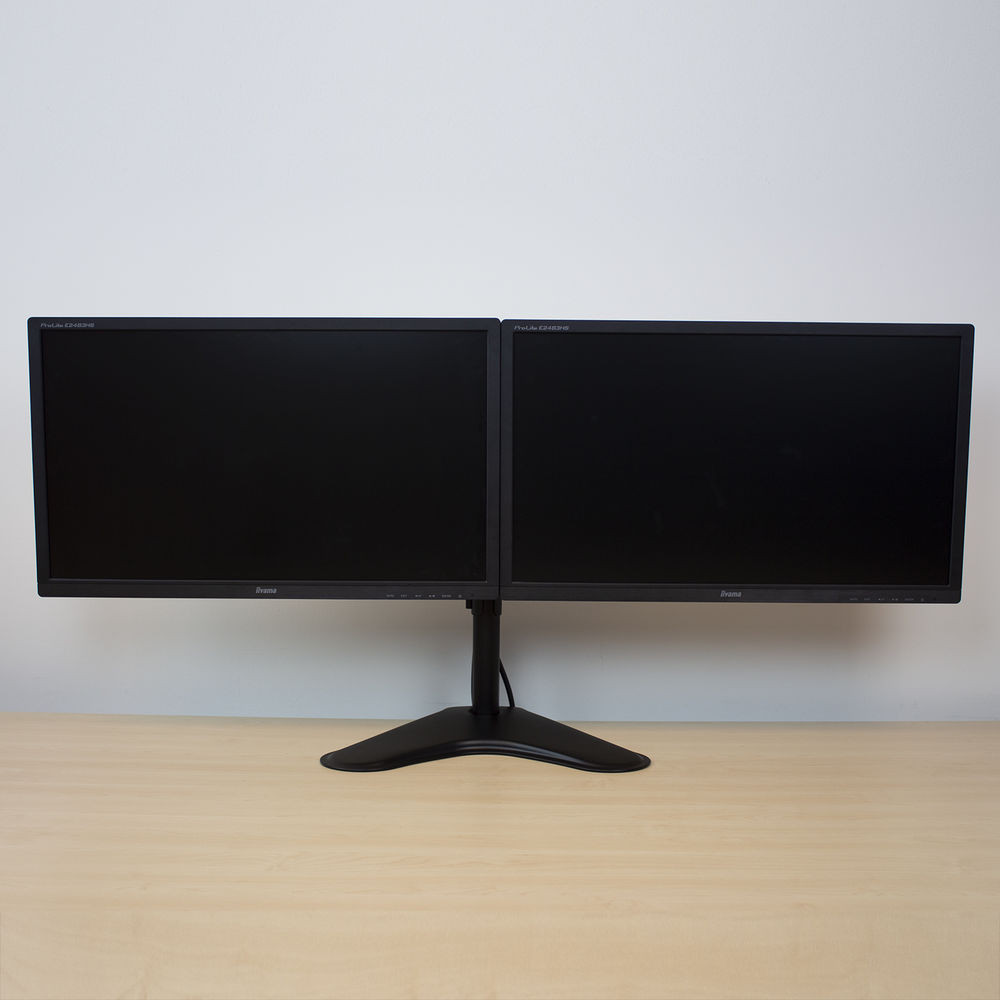 Two black computer monitors on a light wood desk. White wall background.