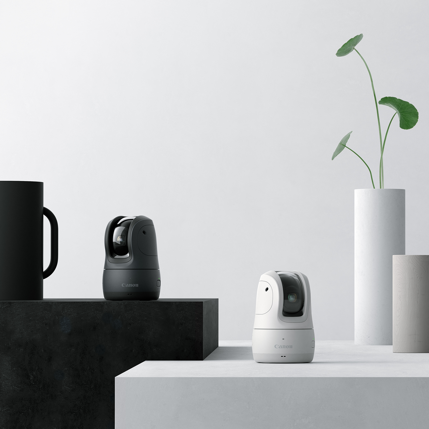 Two Canon security cameras, one black, one white, on a white platform with a black mug and plant.