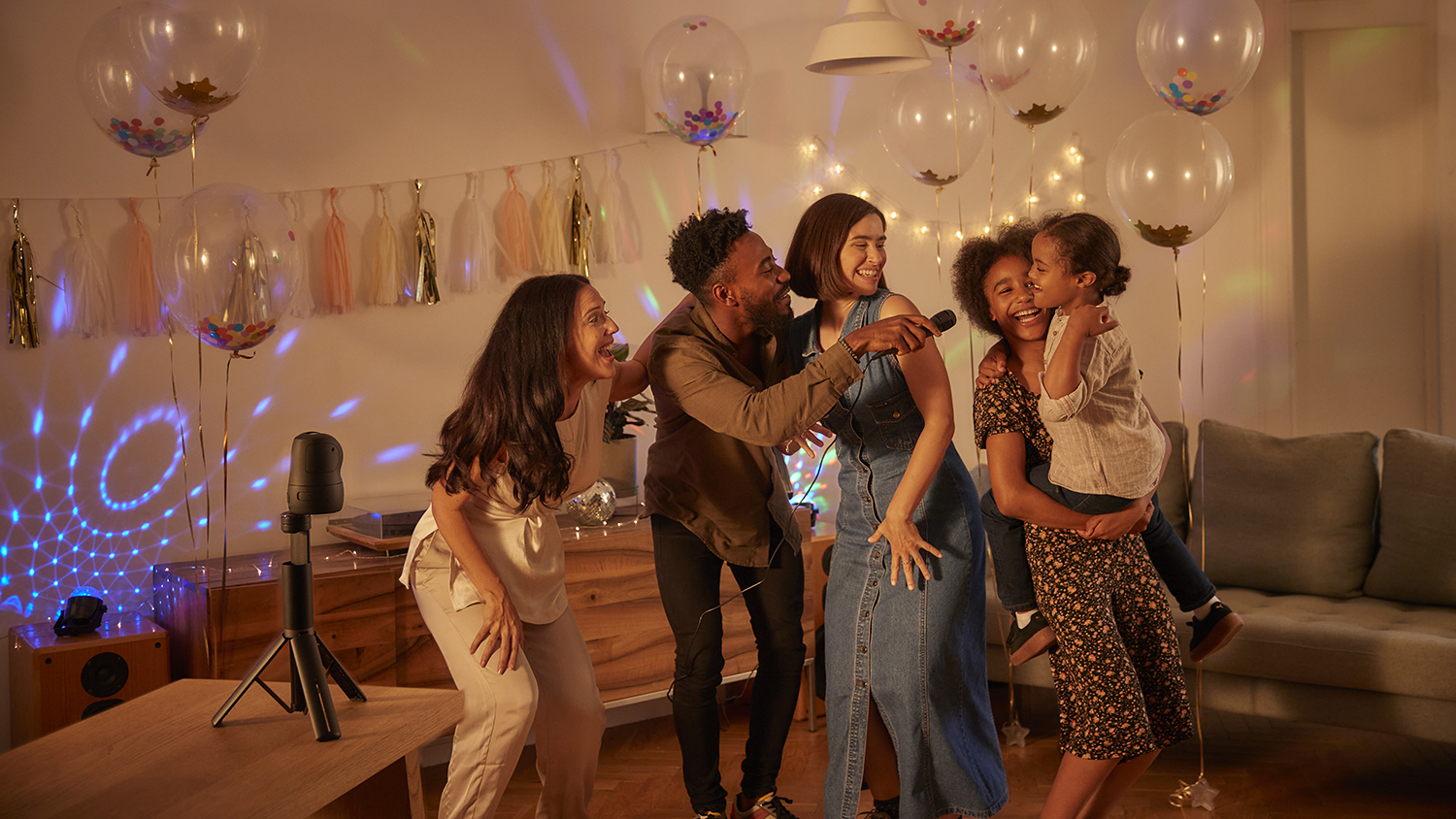 A family singing karaoke in a living room, with balloons and decorative lights in the background.