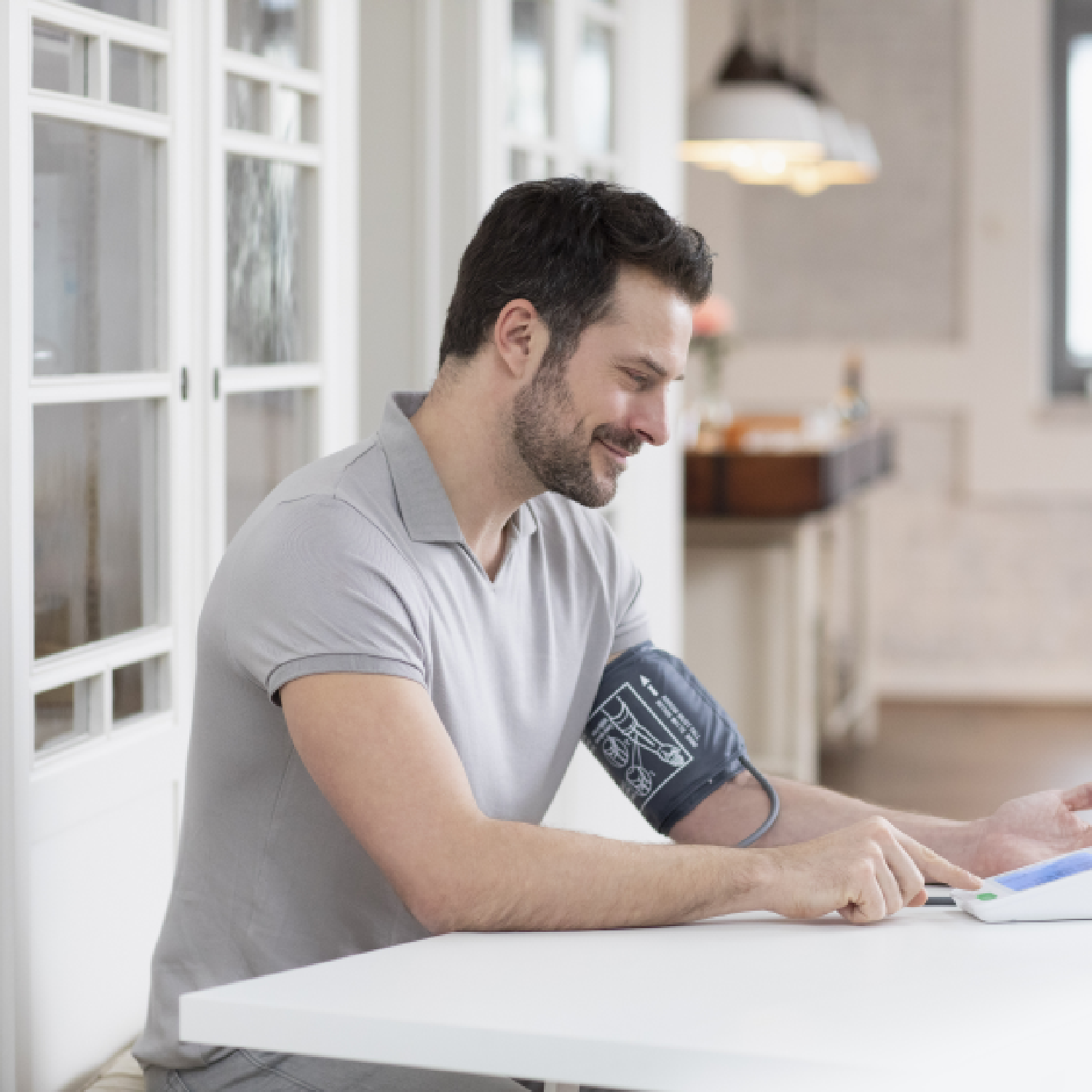 Man using a blood pressure monitor on his arm, sitting at a white table, smiling indoors.