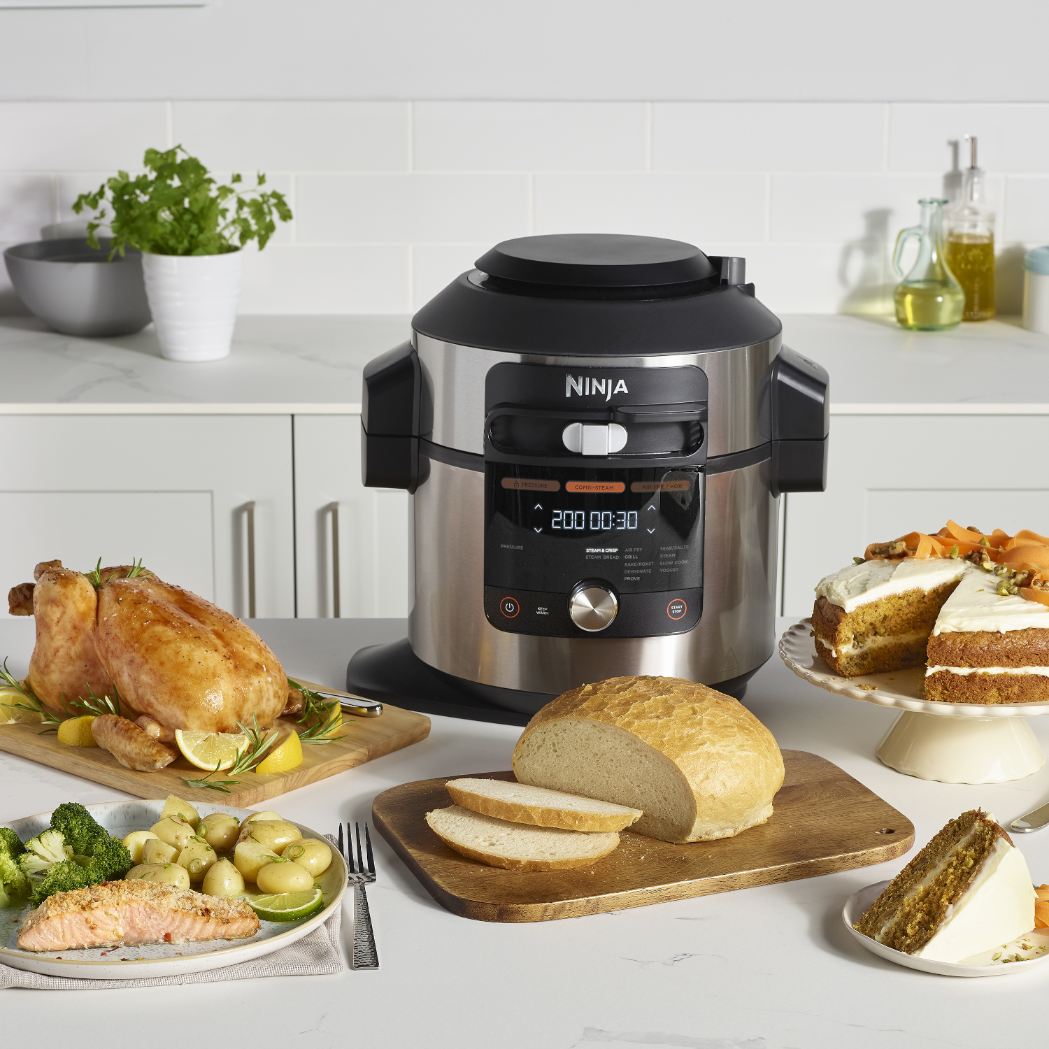 Een Ninja multicooker met gekookt voedsel eromheen: een hele kip, een zalmgerecht, brood en cake.