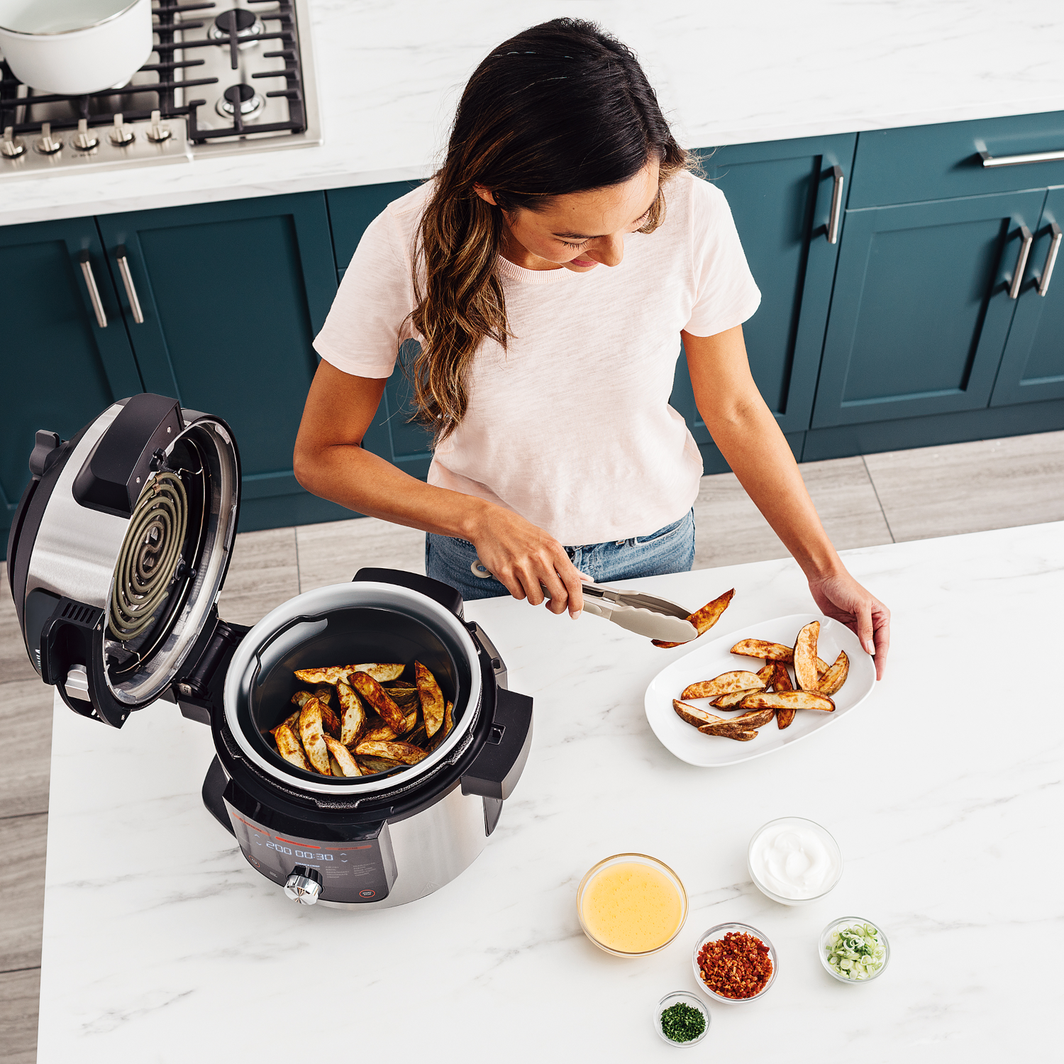 Vrouw gebruikt tang om aardappelpartjes uit de airfryer te halen. Ingrediënten in schalen in de buurt.