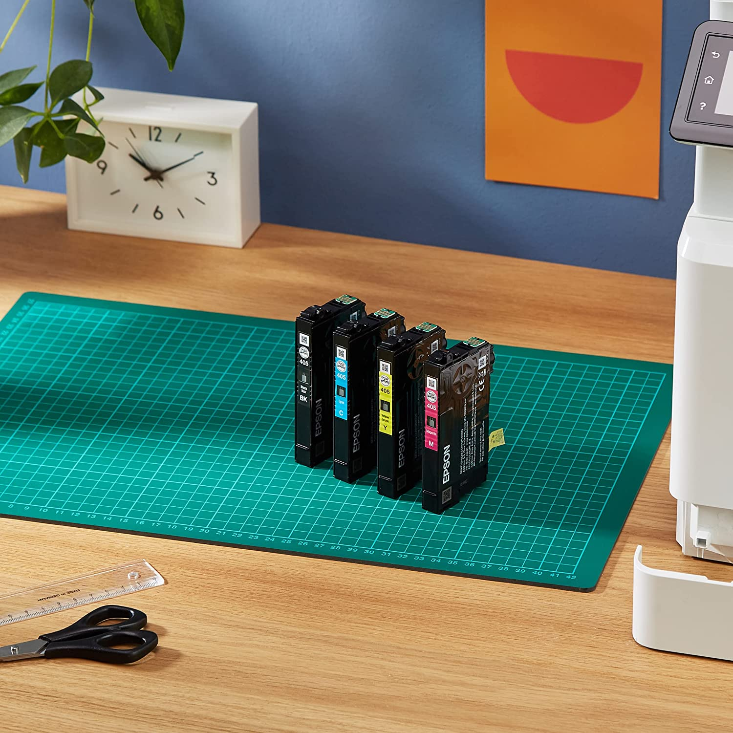 Cartuchos de tinta en una esterilla de corte, con tijeras y una regla. Impresora visible en el fondo. Mesa de madera.