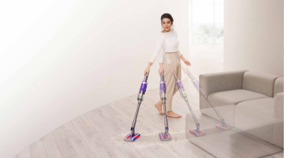 Una mujer aspirando el suelo con una aspiradora inalámbrica. Interior blanco. Suelo de madera gris.