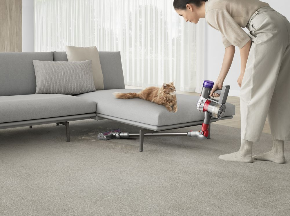 Mujer aspirando el suelo junto a un gato en un sofá gris con una aspiradora.