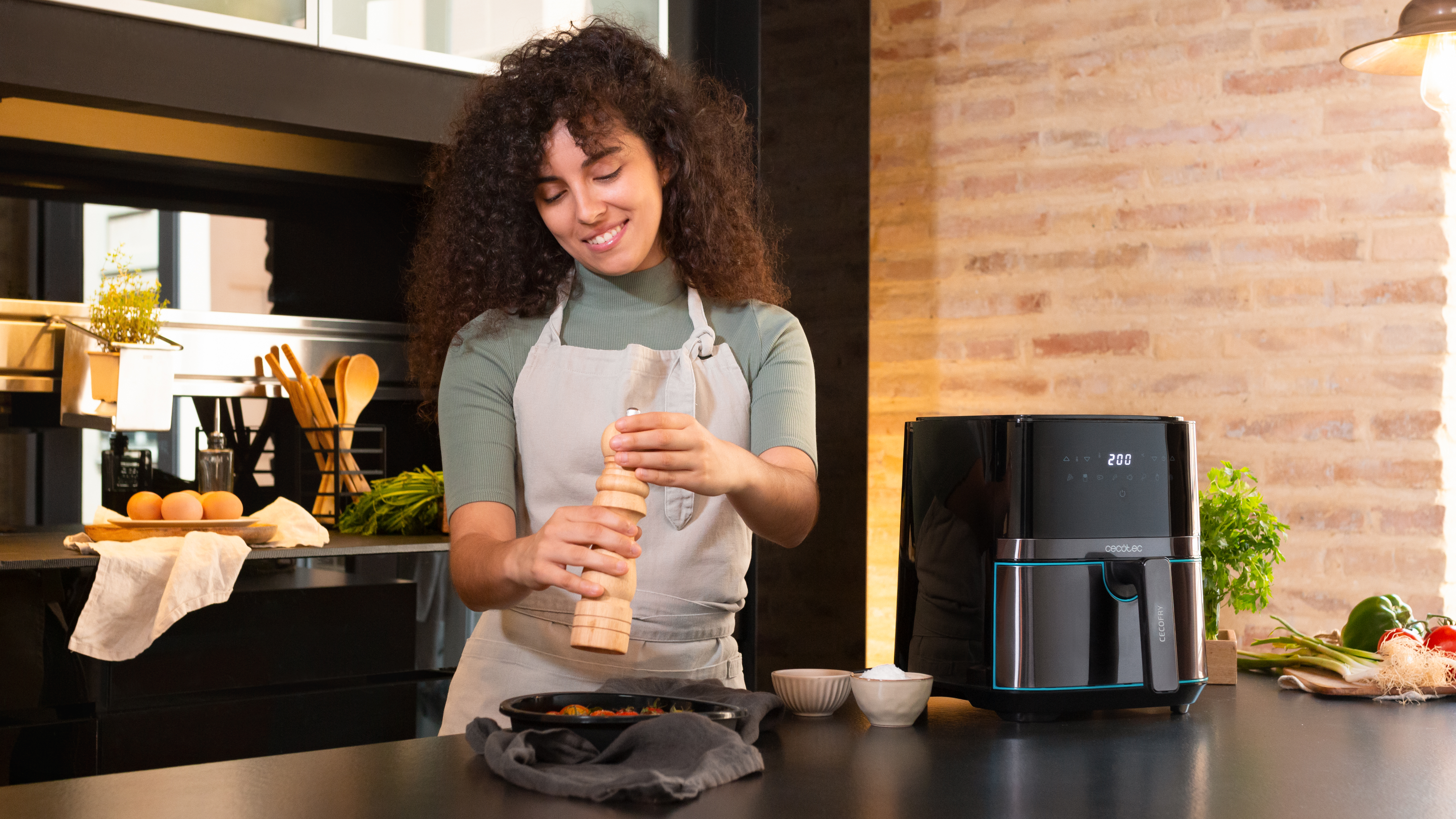 Mujer en cocina usa molinillo de pimienta. Freidora de aire negra en encimera. Pared de ladrillo al fondo.