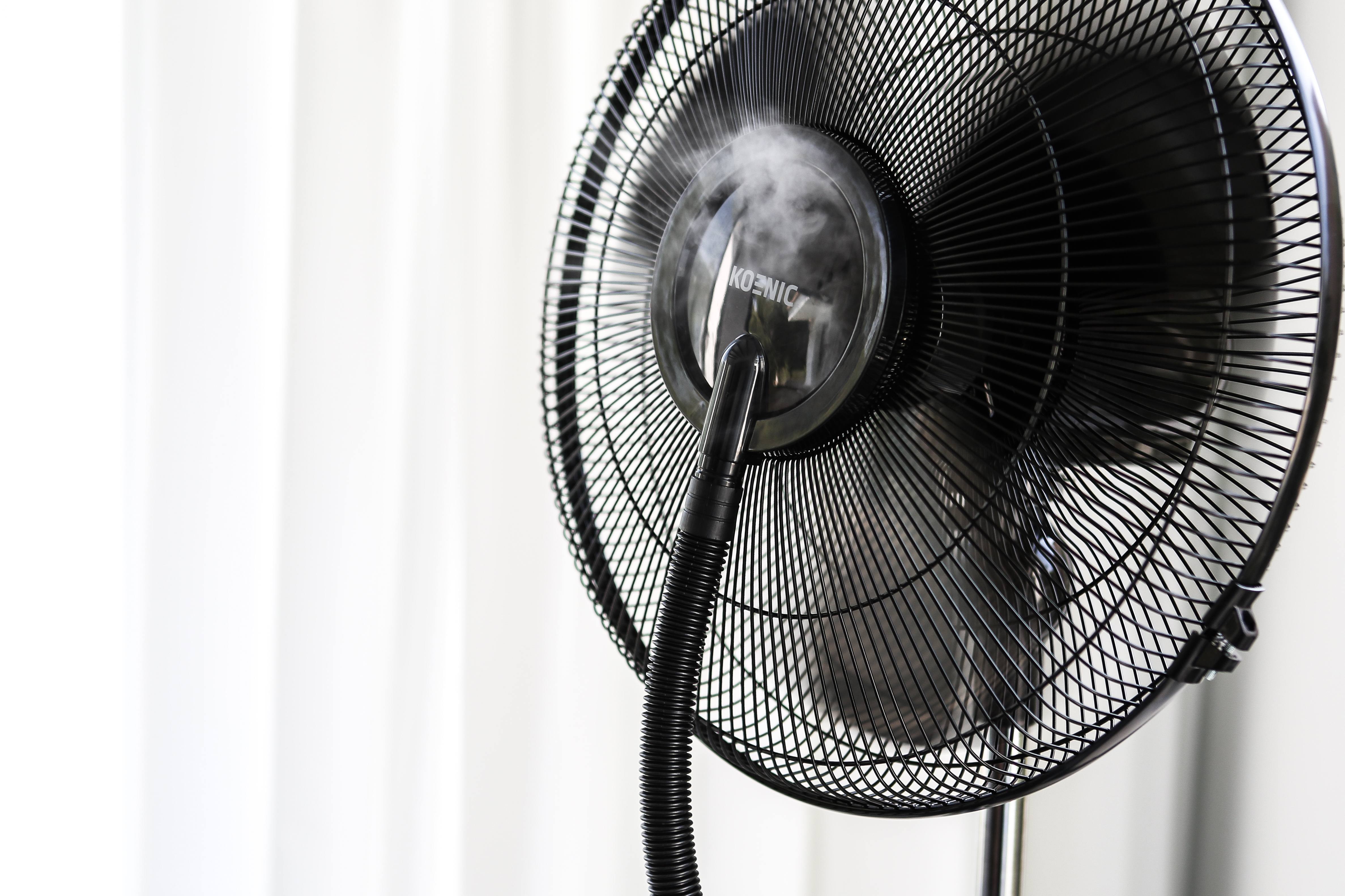 Schwarzer Standventilator mit Nebel. Lüfterflügel sind sichtbar. Weiße Vorhänge im Hintergrund. Der Ventilator ist an.