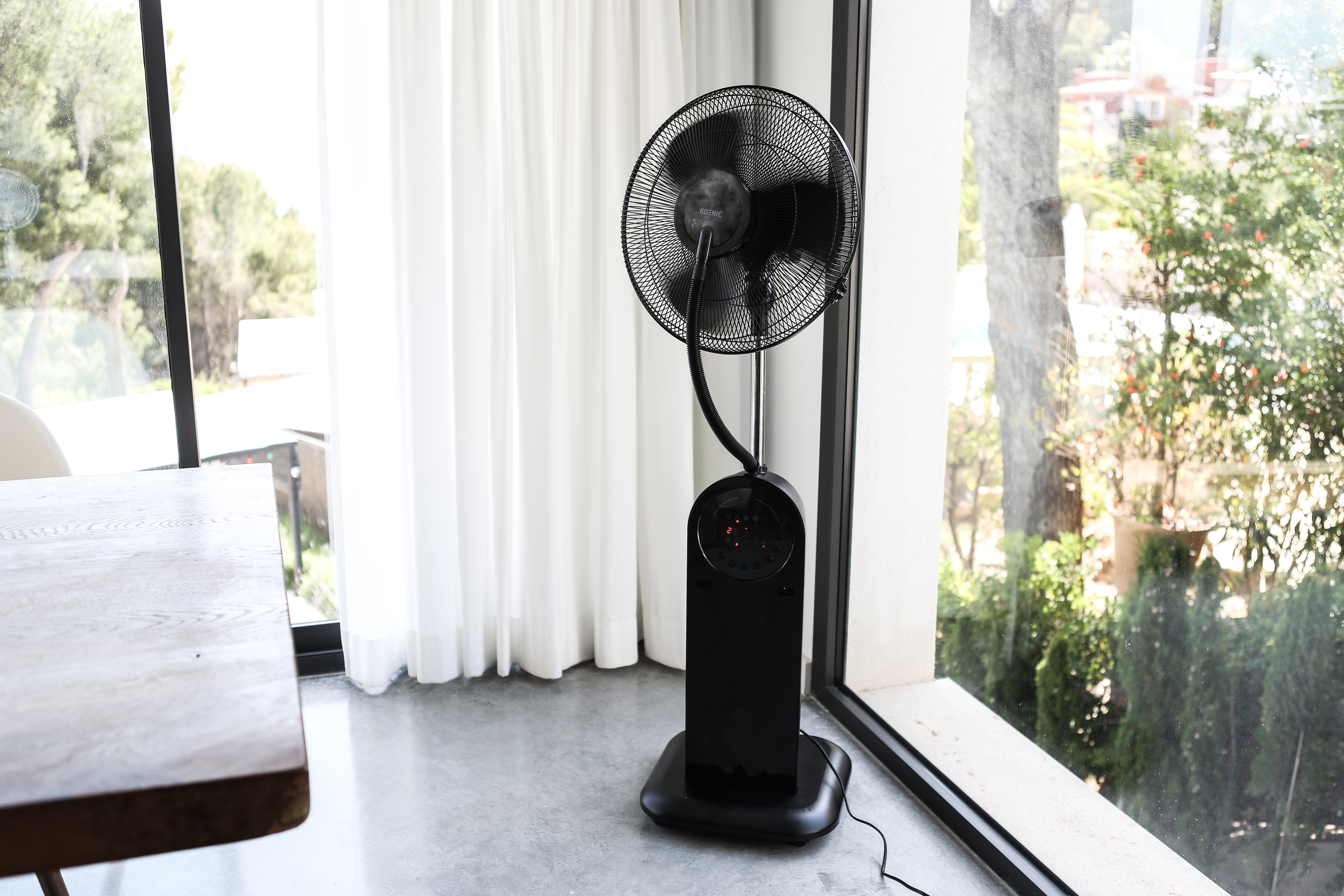 Schwarzer Standventilator. Der Ventilator steht vor einem Fenster mit Bäumen.
