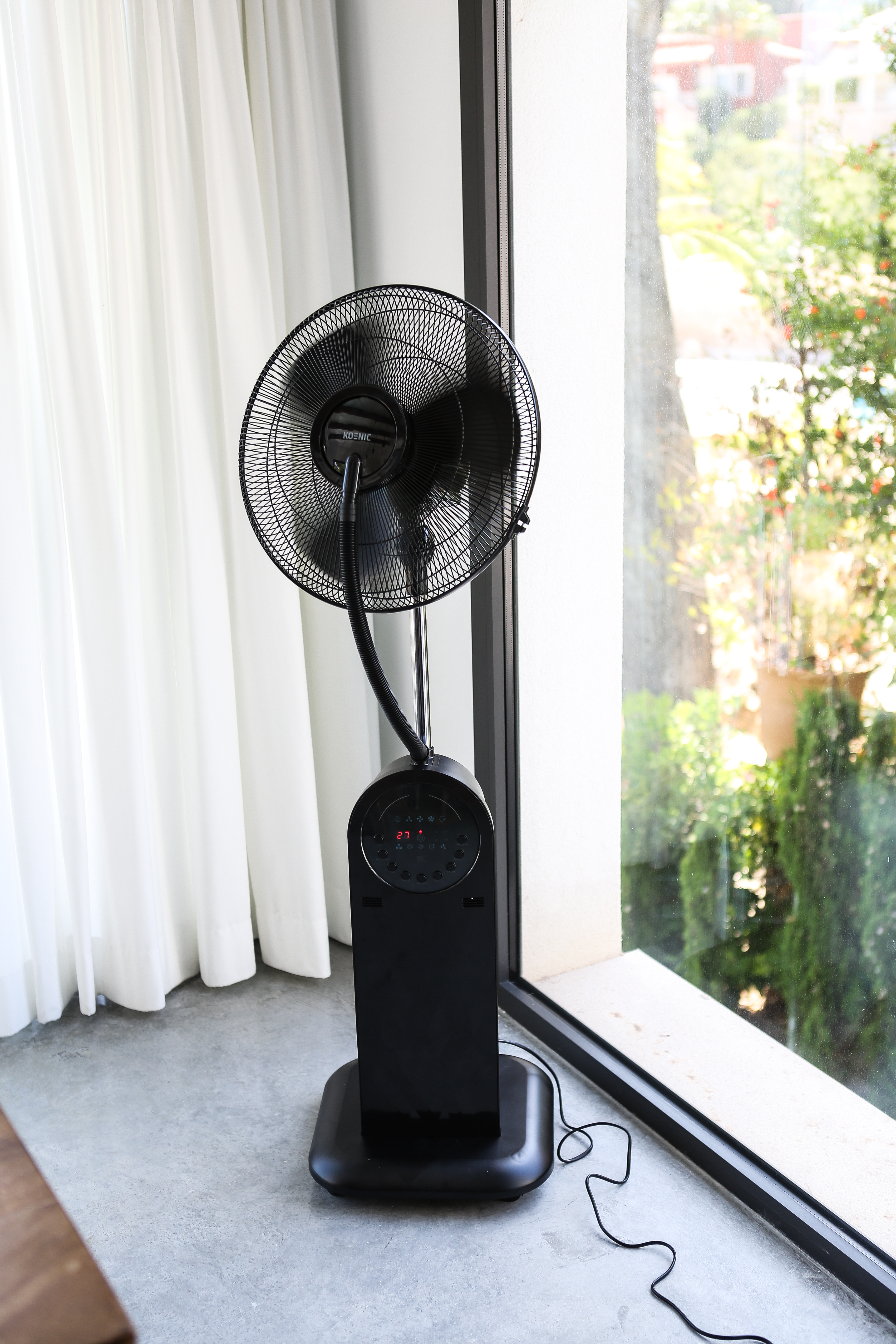 Schwarzer Ventilator auf Ständer am Fenster, weiße Vorhänge und Außenansicht.