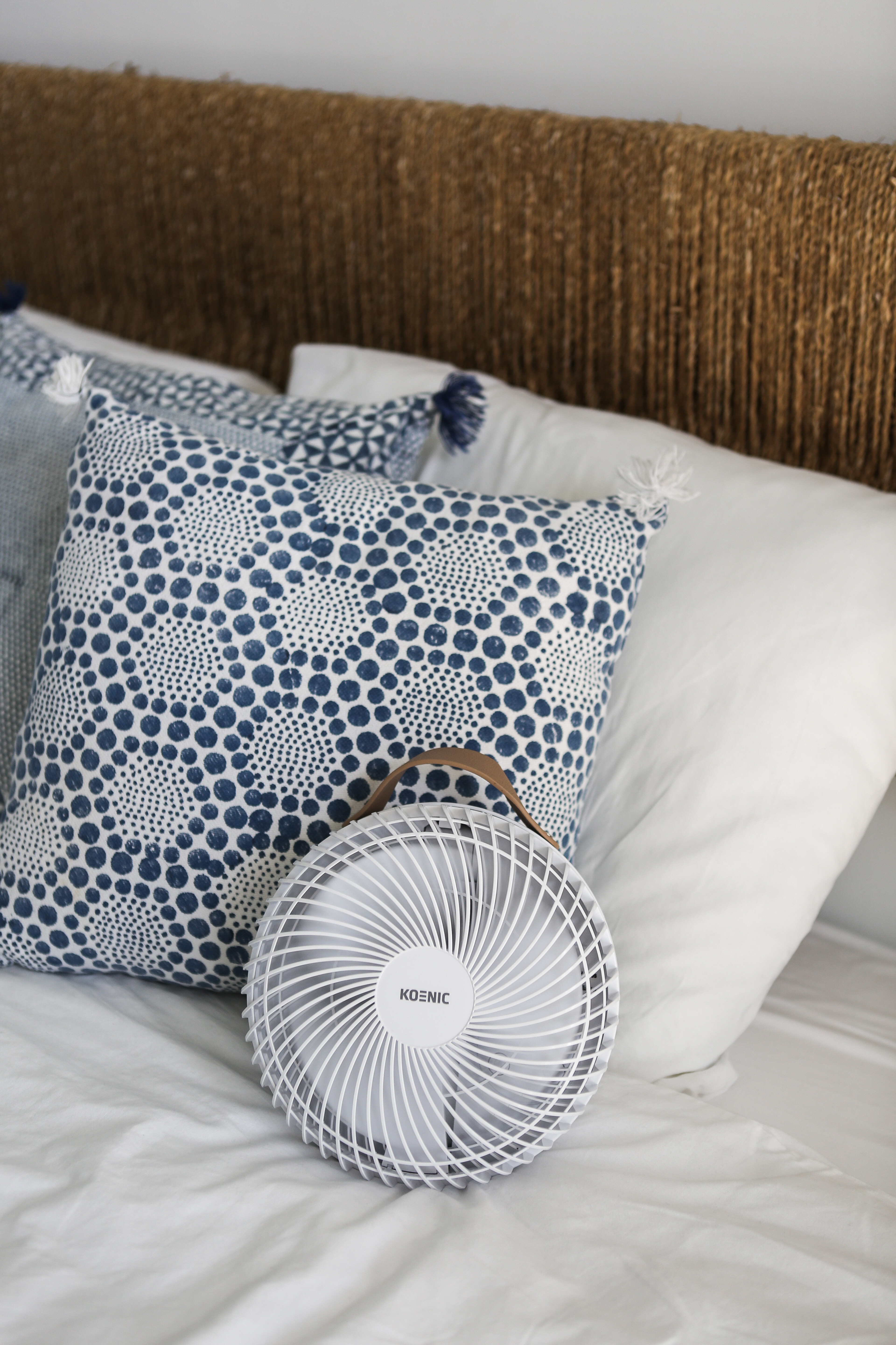 Ein kleiner weißer Ventilator auf einem Bett mit Kissen und einem gewebten Kopfteil.