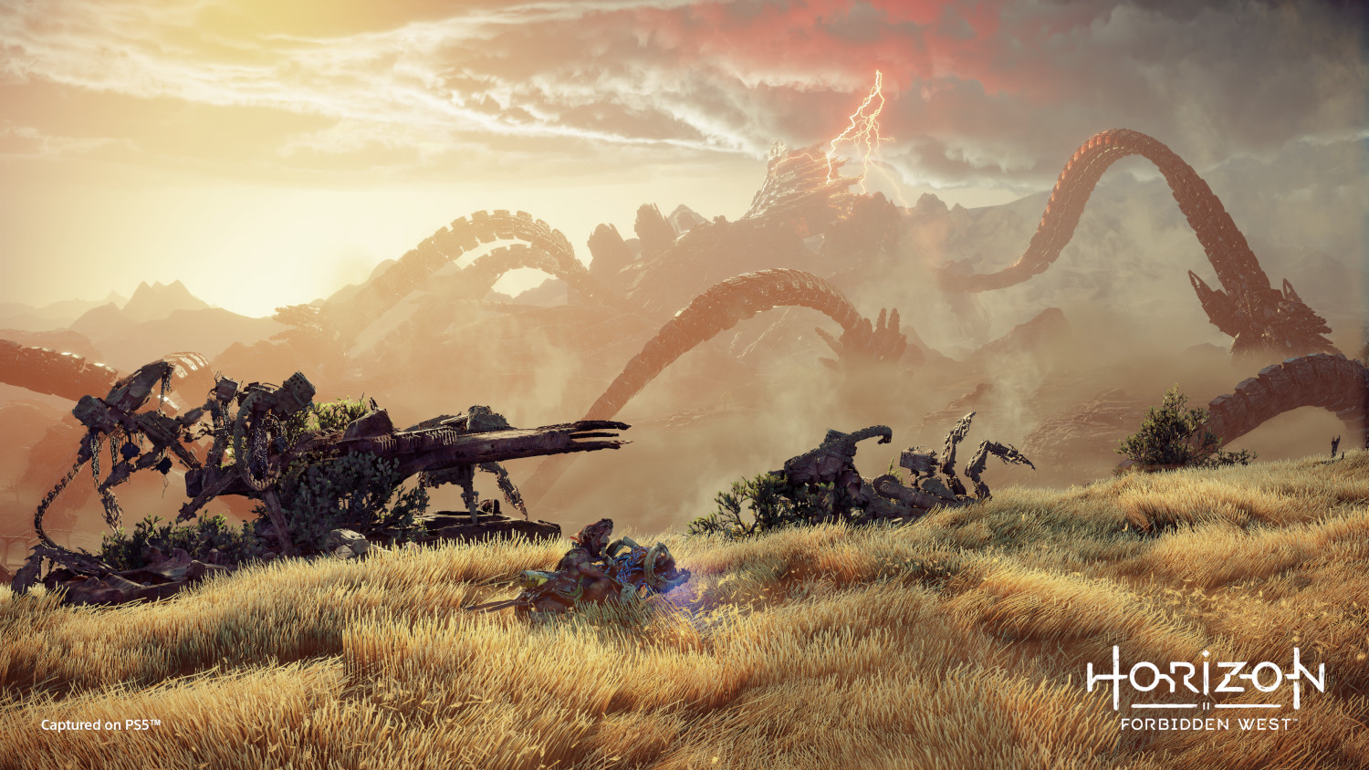 A player character crouches in tall grass near robotic machines. Giant mechanical serpent tails rise in the background.
