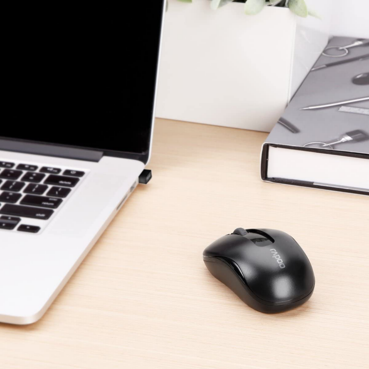 Ratón inalámbrico negro junto a un portátil y un libro sobre un escritorio de madera.