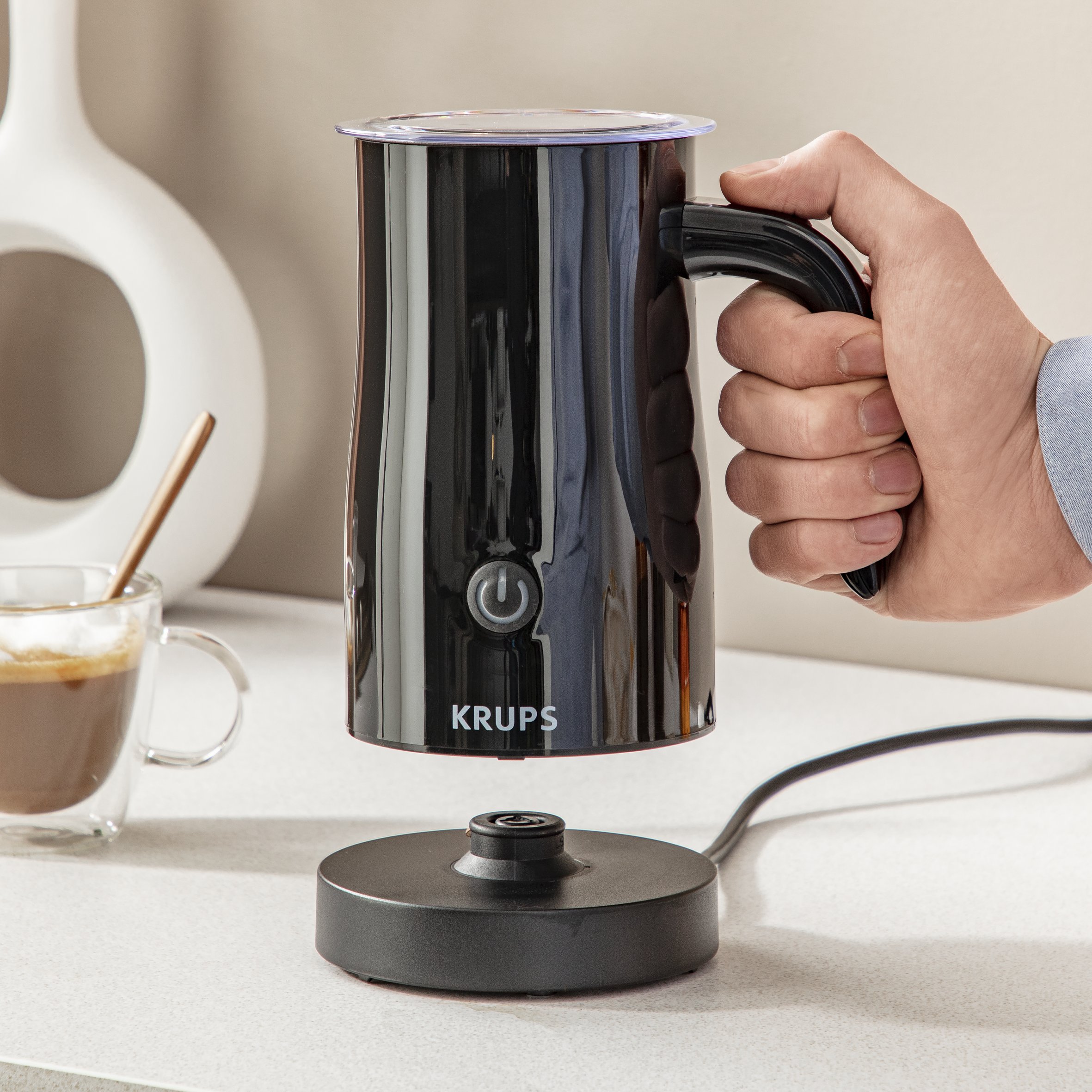 Una persona sosteniendo un espumador de leche Krups negro, con un vaso de café al lado.