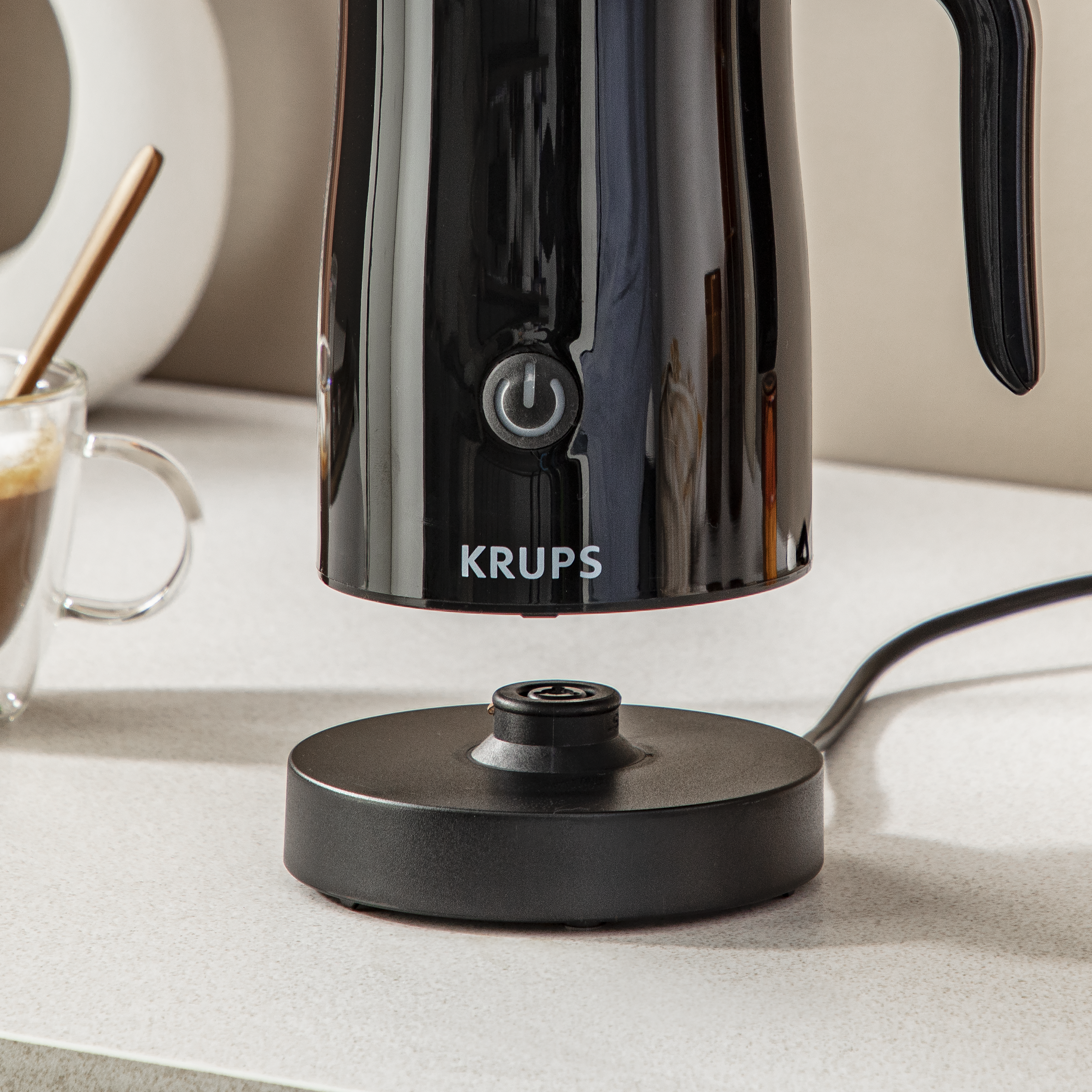 Un espumador de leche Krups negro con un botón de encendido. Un vaso de café está en el fondo.