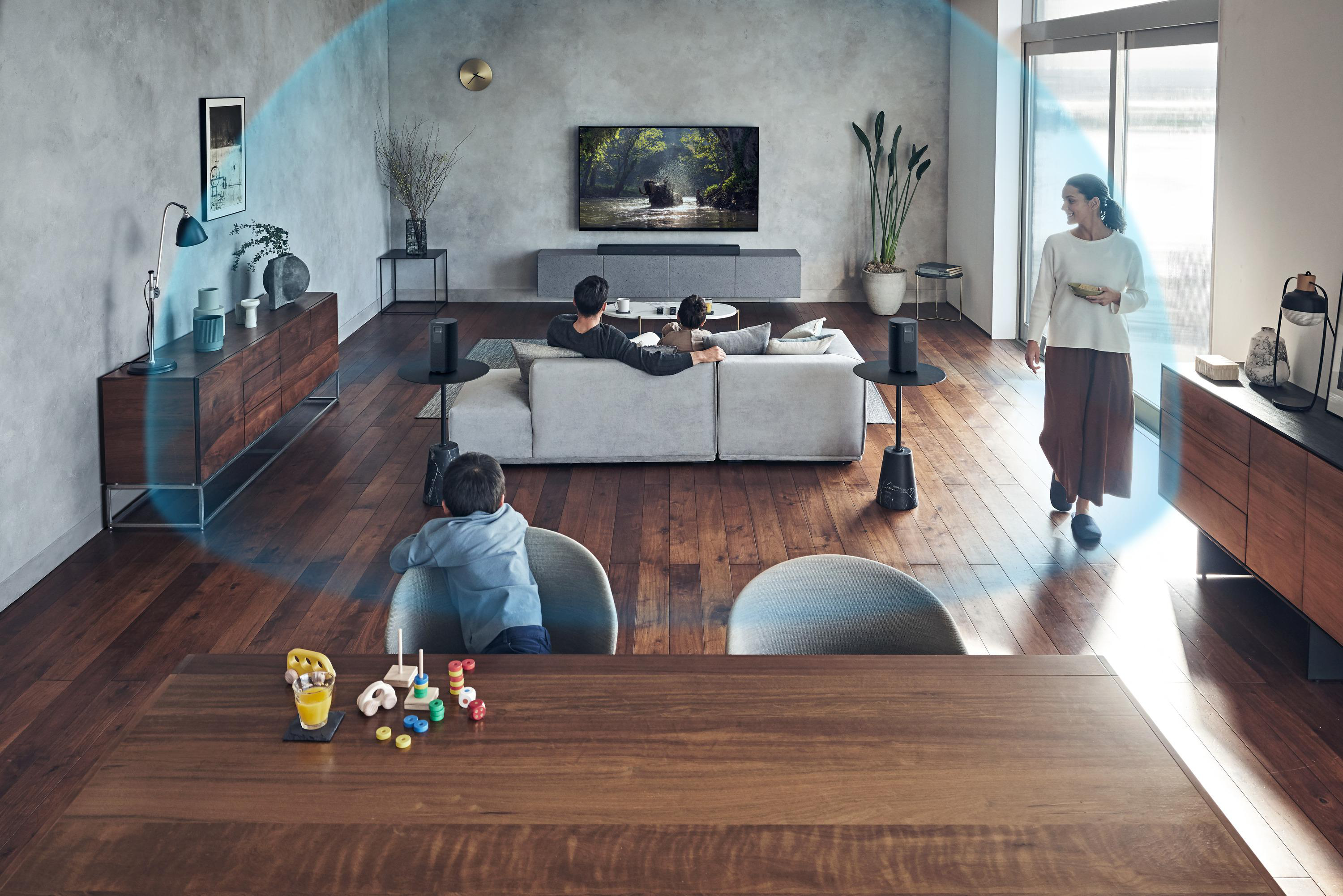 Wohnzimmer mit Menschen, Fernseher, Sofa, Lautsprechern und Esstisch.