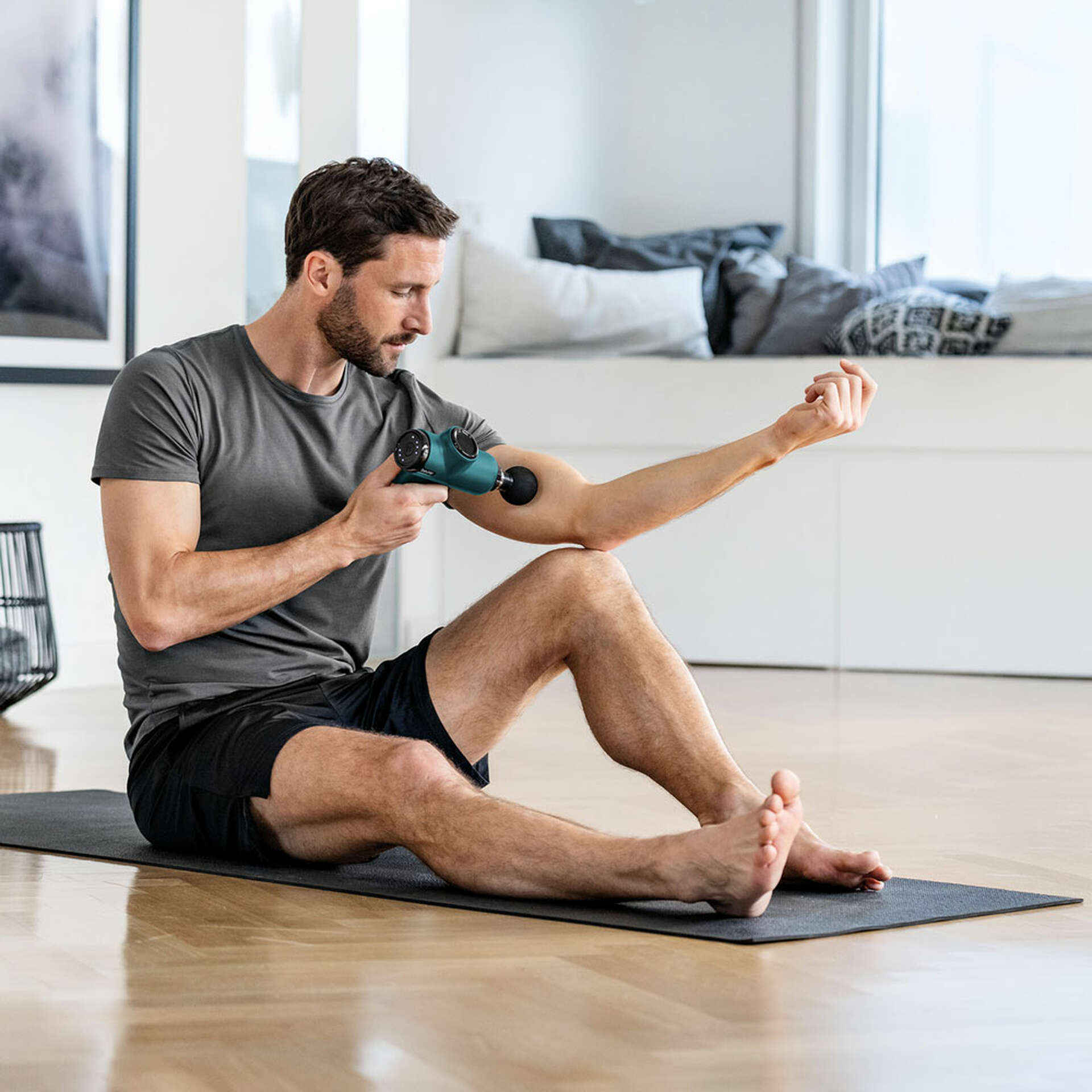 Ein Mann benutzt eine Massagepistole an seinem Arm und sitzt auf einer Matte. Ein Fenster und ein Bett sind im Hintergrund.