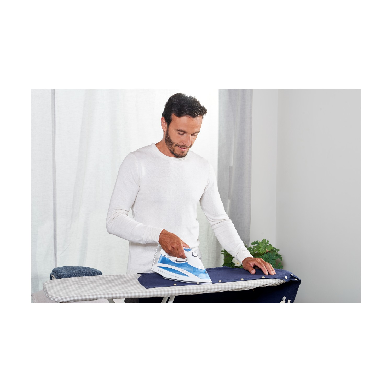 Un hombre planchando una camisa azul en una tabla de planchar. Fondo blanco.